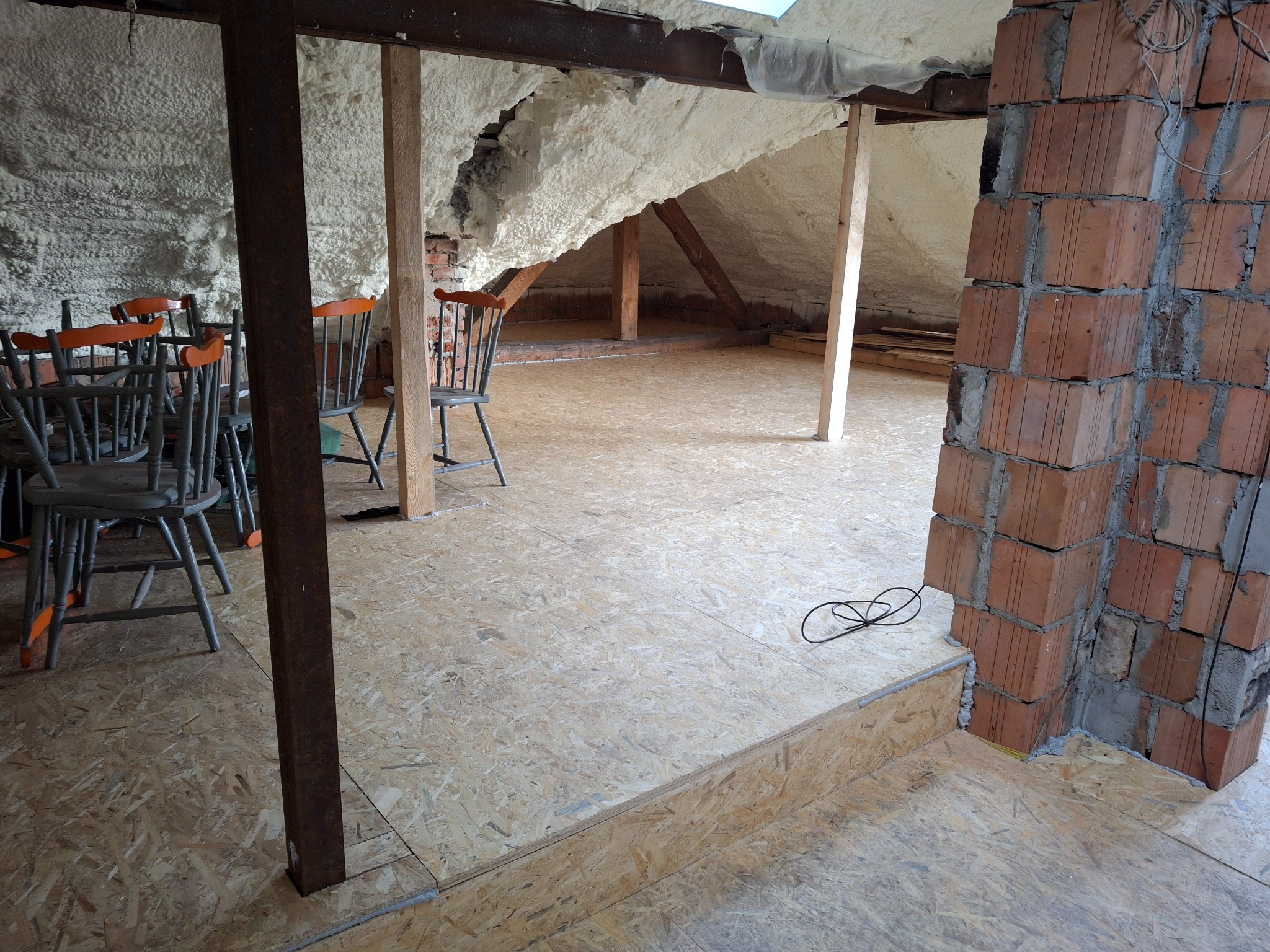 Poddasze w trakcie adaptacji: ocieplone pianką ściany, surowa cegła komina i podłoga z płyt OSB, widoczne drewniane podpory i krzesła.