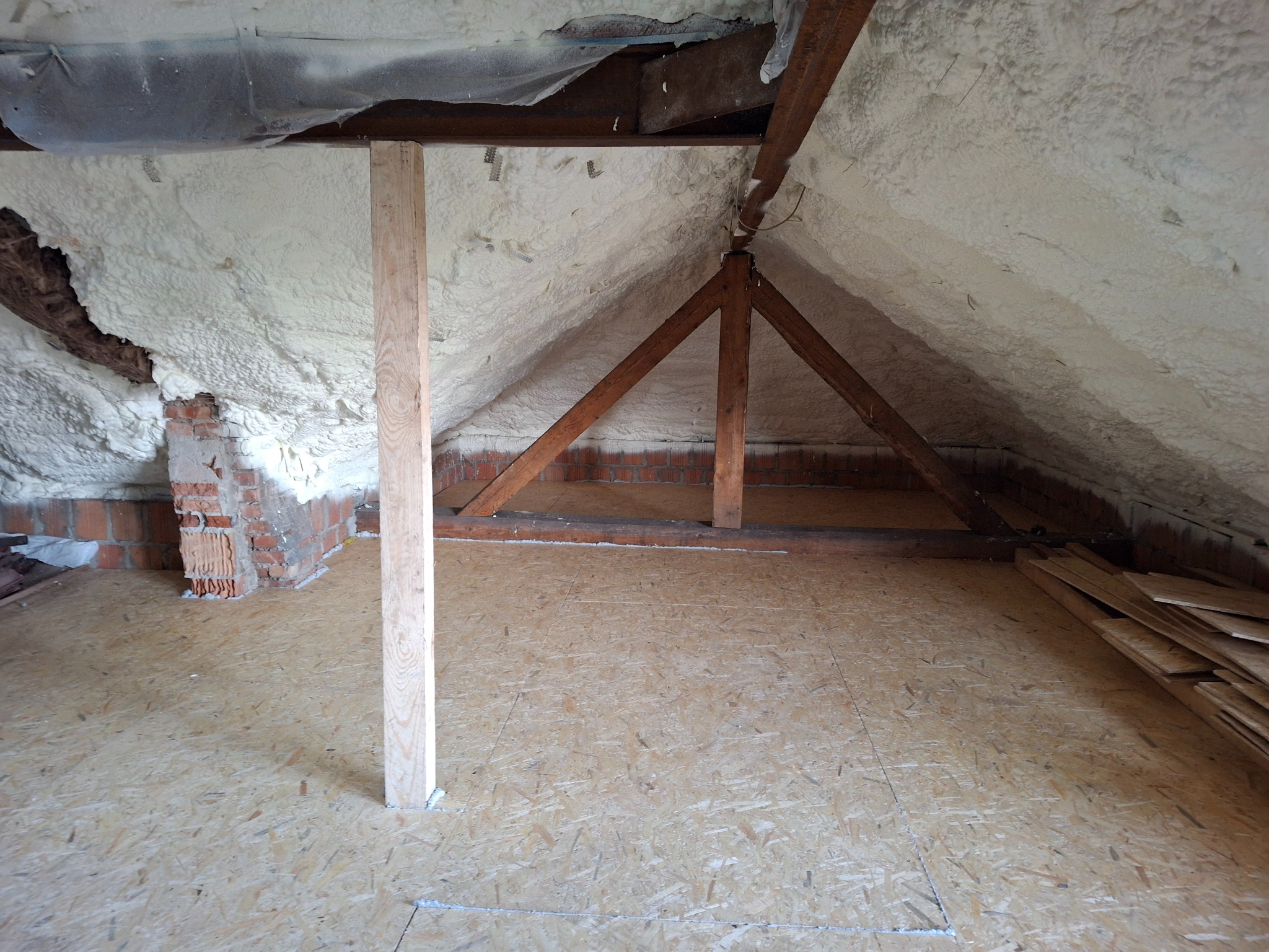 Poddasze z ociepleniem pianką PUR, widoczne drewniane elementy konstrukcji dachu i podłoga z płyt OSB, fragment muru z cegły.