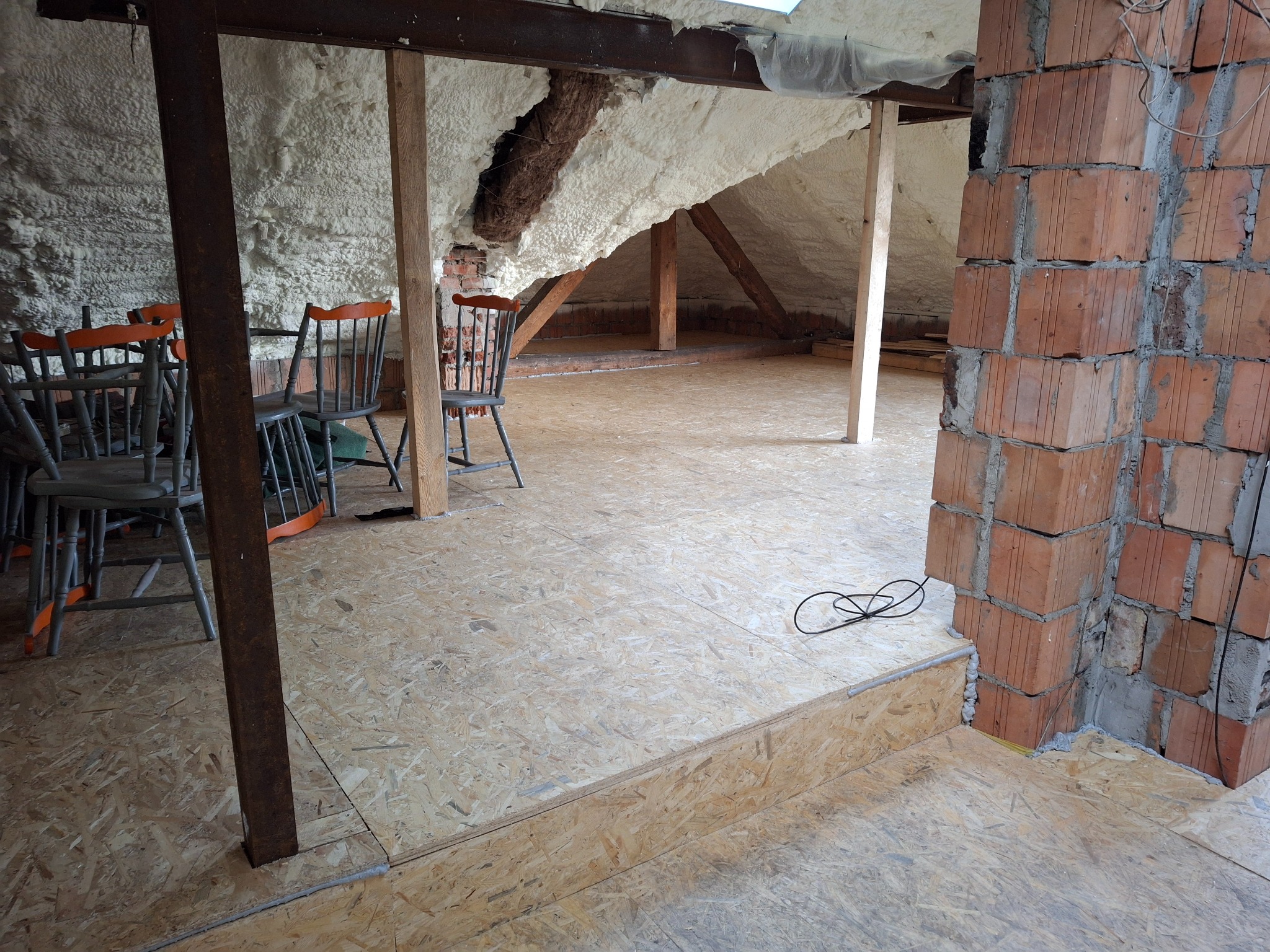 Poddasze w trakcie adaptacji: izolacja pianą, podłoga z płyt OSB, fragment murowanego komina, sterta krzeseł z pomarańczowymi oparciami w tle.