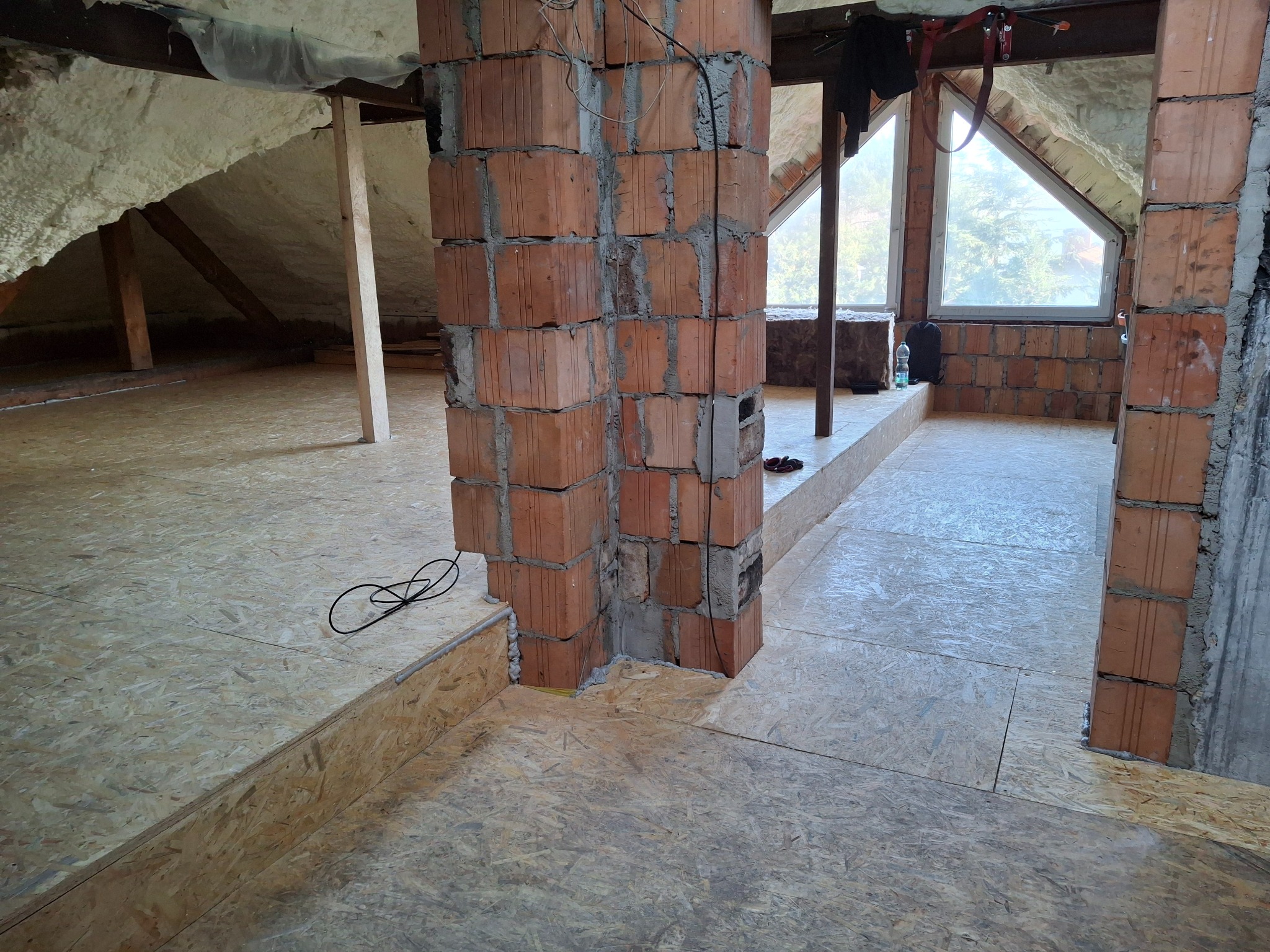 Poddasze w trakcie adaptacji: surowa ceglana kolumna, ocieplone pianą ściany skośne, podłoga z płyt OSB, okno dachowe trójkątne. Widoczne przewody elektryczne i buty.