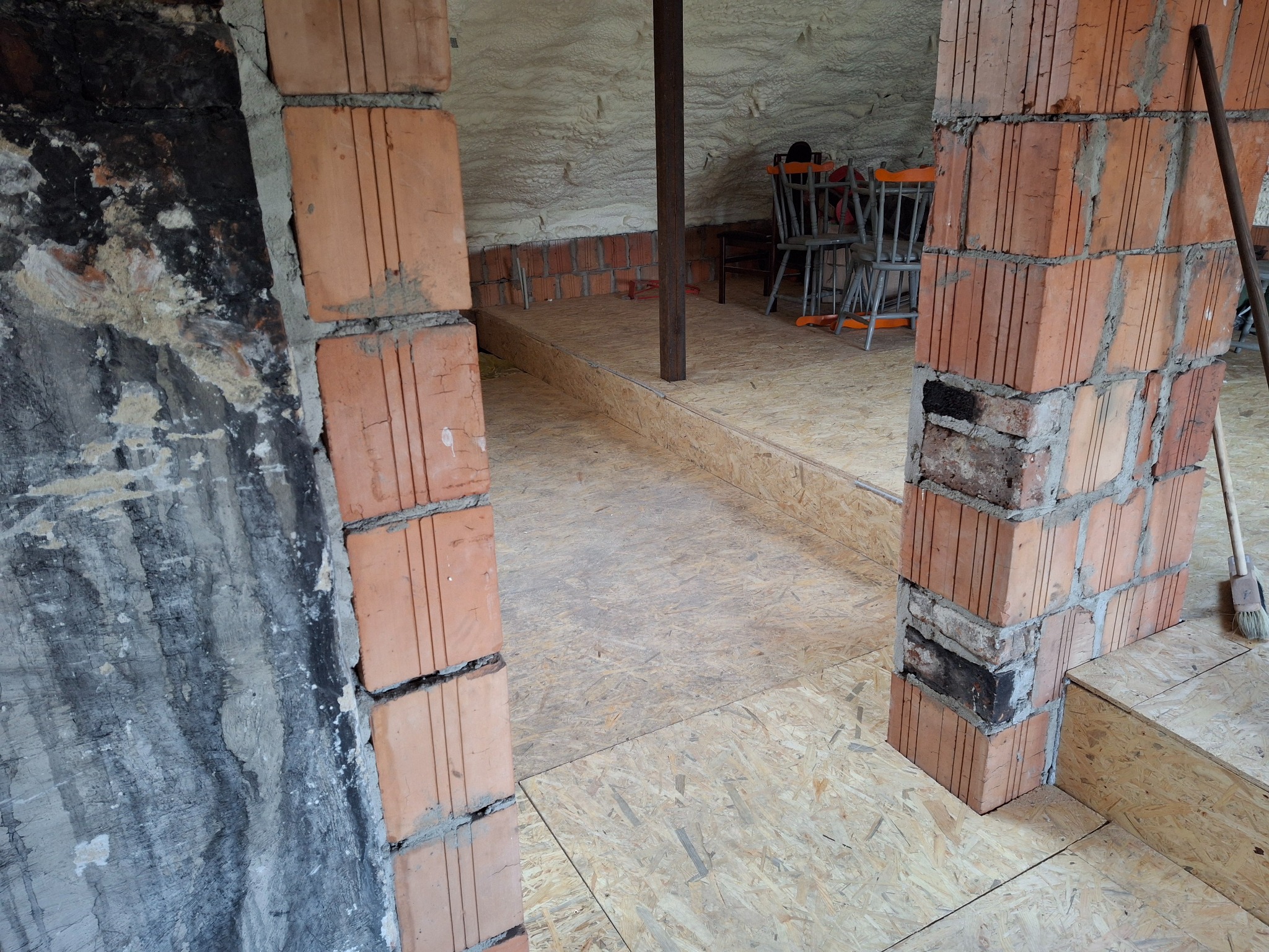 Wnętrze poddasza w trakcie adaptacji: surowa ściana z cegły, płyta OSB na podłodze, ocieplenie pianką na skośnym suficie, widoczne krzesła w tle, ślady pożaru na ścianie z lewej strony.