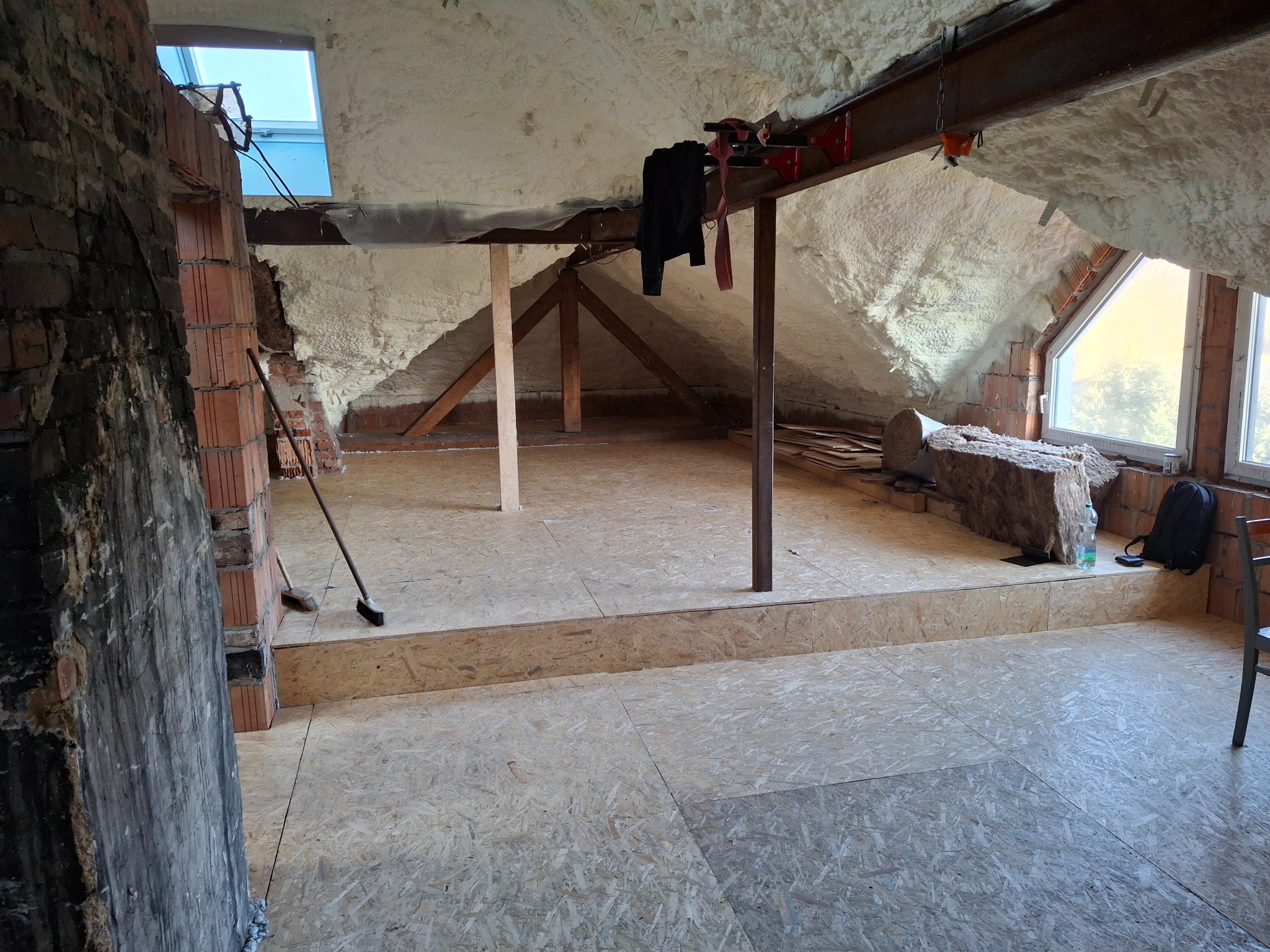 Poddasze w trakcie adaptacji, z widoczną izolacją piankową na skosach dachu, podłogą z płyt OSB na różnych poziomach, fragmentami muru z cegły, oknem dachowym i roboczym bałaganem.