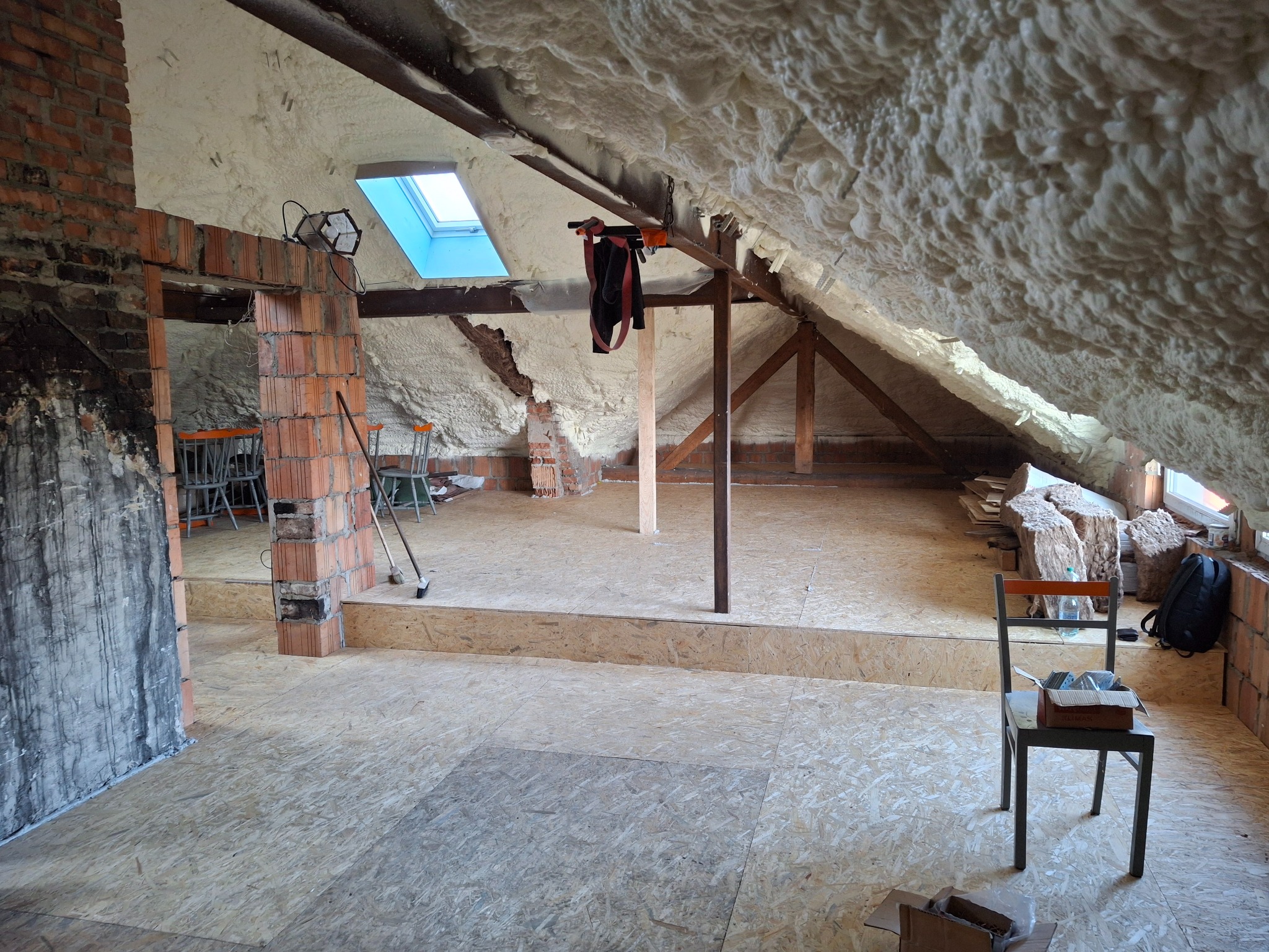 Poddasze w trakcie ocieplania pianą PUR, widoczne fragmenty muru z cegły, okno dachowe i tymczasowe oświetlenie