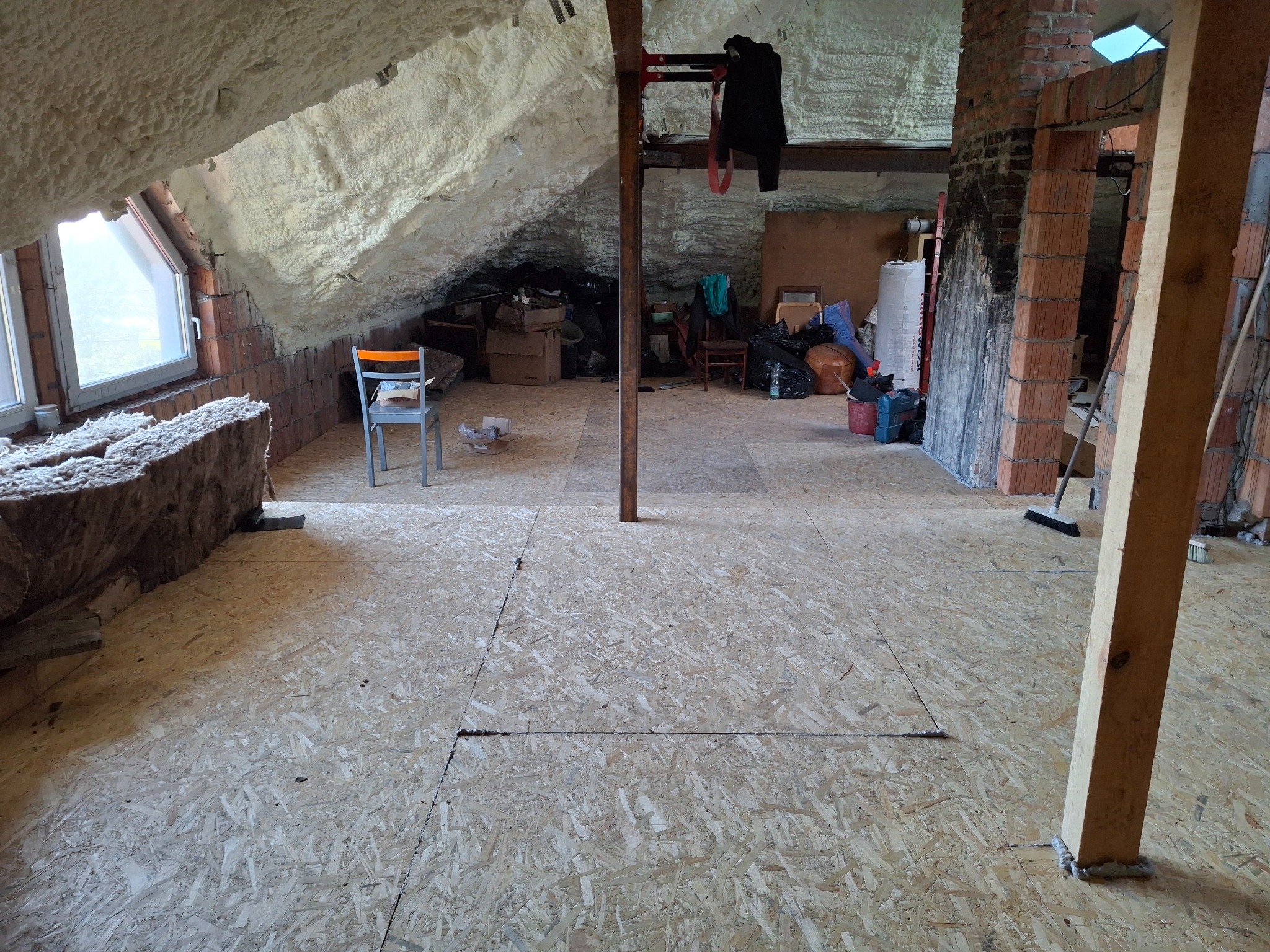 Poddasze w trakcie adaptacji: ocieplone skosy z pianki, surowa podłoga z płyt OSB, widoczne elementy konstrukcyjne i materiały budowlane, przygotowane do dalszych prac wykończeniowych.