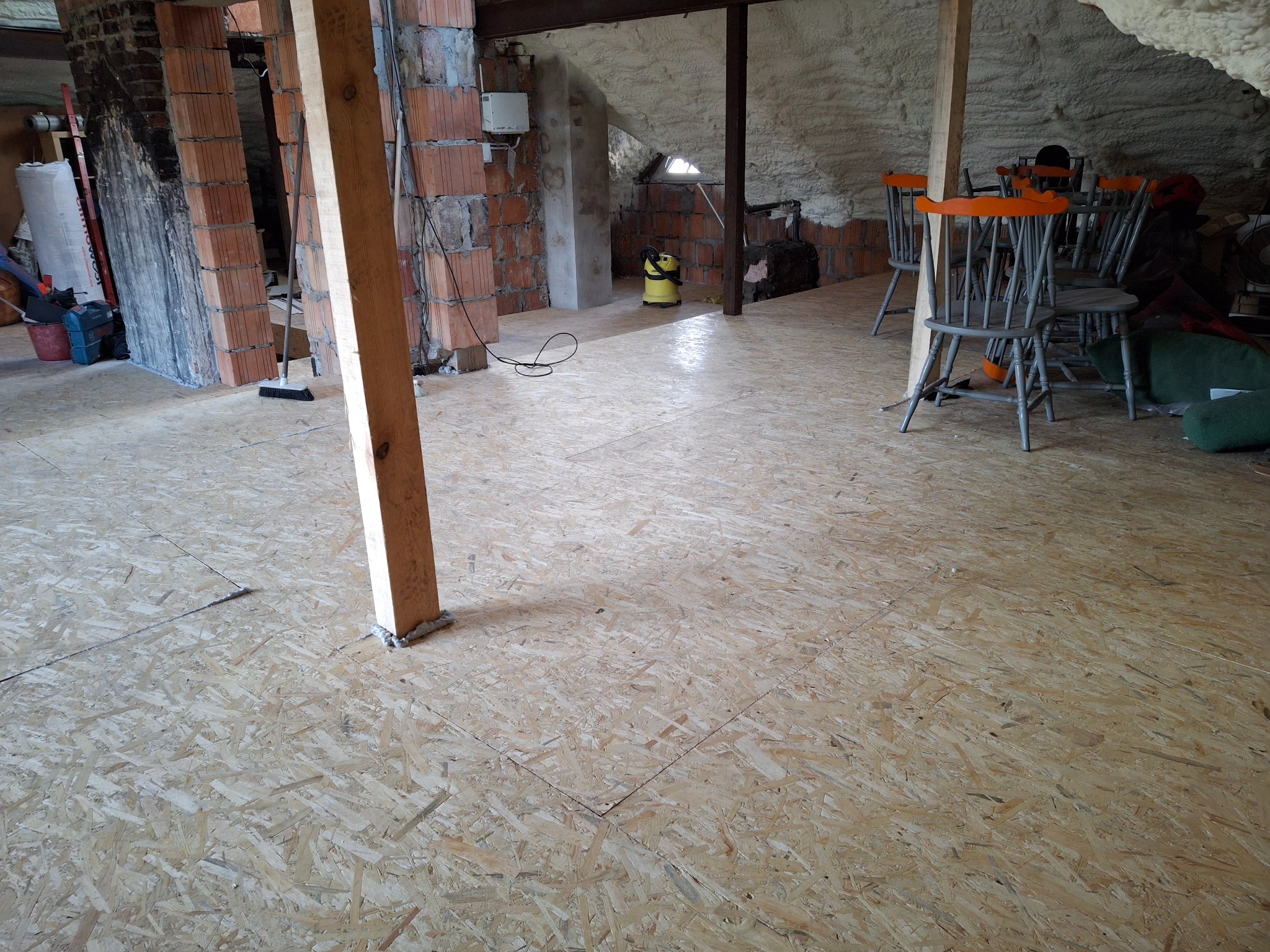 Płyta OSB ułożona na poddaszu, widoczne ściany z cegły, ocieplenie pianką, krzesła w tle i odkurzacz przemysłowy, etap prac wykończeniowych.