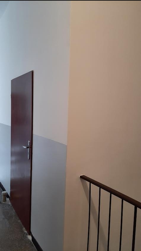 Świeżo pomalowane ściany w odcieniach bieli i beżu, widoczna odnowiona, ciemna stolarka drzwiowa i fragment balustrady schodowej. Minimalistyczne wnętrze po renowacji.