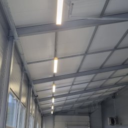 Kacper Gubała Instalacje Elektryczne - Wnętrze hali warsztatowej z rzędem podłużnych lamp LED zamontowanych pod sufitem, widoczne kanały serwisowe i pracownicy w oddali.