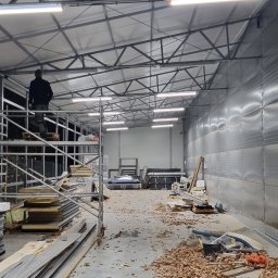 Kacper Gubała Instalacje Elektryczne - Wnętrze hali w trakcie prac wykończeniowych; widoczne rusztowanie z pracownikiem, zamontowane lampy LED, materiały budowlane i rozsypane liście na podłodze.