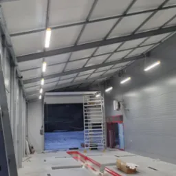 Wnętrze hali warsztatowej z surowym betonem, oświetleniem LED, kanałami naprawczymi z czerwonymi akcentami i rozstawionym rusztowaniem, widoczne okablowanie i narzędzia.