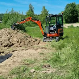 Pomarańczowa mini-koparka Kubota KX018-4 nabiera ziemię z pryzmy na trawiastym terenie w słoneczny dzień.