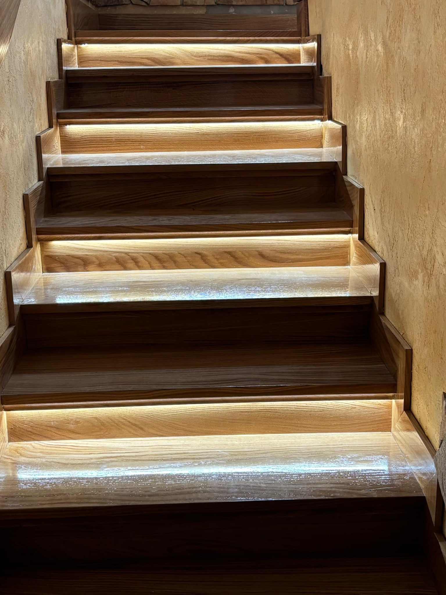 Drewniane schody z podświetleniem LED na każdym stopniu, ciepłe światło odbija się od polerowanej powierzchni stopni, obok ściana o fakturze tynku.