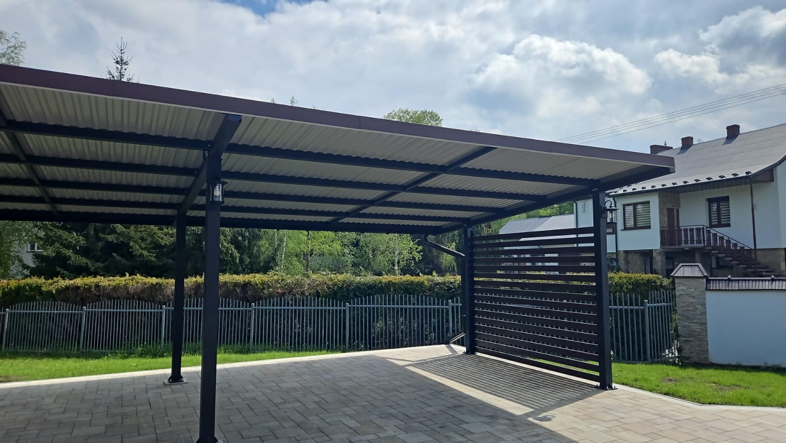 Zadaszenie w kolorze grafitowym z oświetleniem zewnętrznym w stylu retro, przylegające do domu z widocznym fragmentem ogrodu i ogrodzenia.