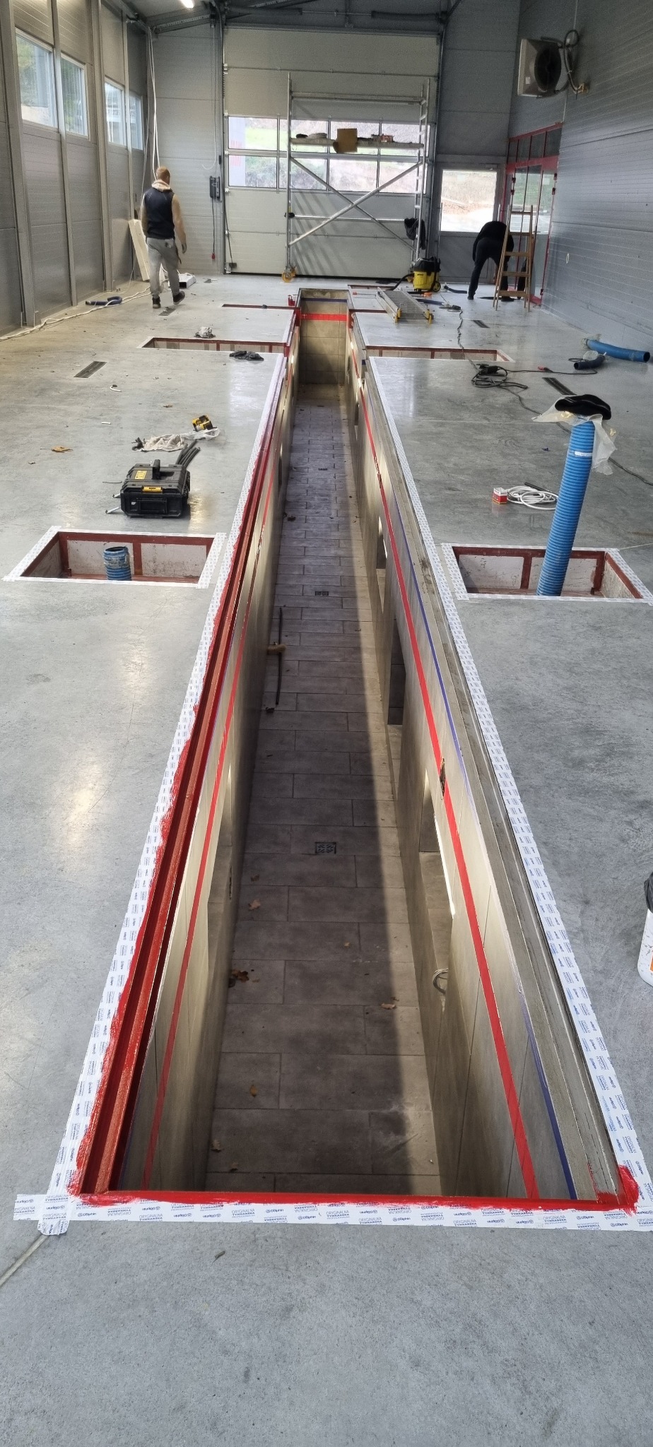 Wnętrze hali warsztatowej z kanałem rewizyjnym w trakcie budowy lub remontu, obrzeża kanału zabezpieczone taśmą malarską i pomalowane na czerwono, widoczne przewody i narzędzia, dwóch pracowników...