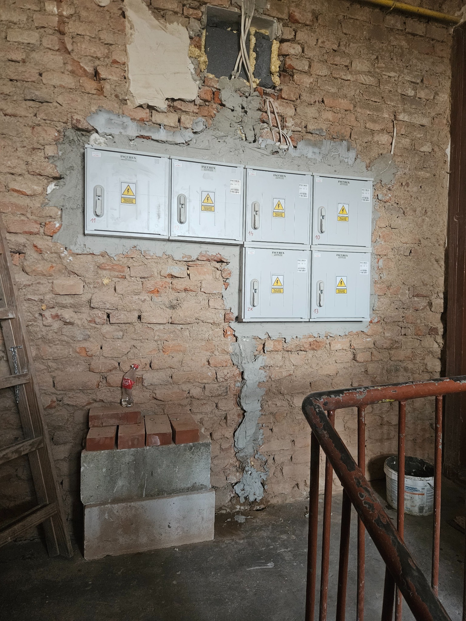 Sześć szarych skrzynek elektrycznych INCOBEX zamontowanych na surowej ceglanej ścianie z widocznymi przewodami i ubytkami tynku, butelka po napoju na cegłach, drabina i barierka w tle.