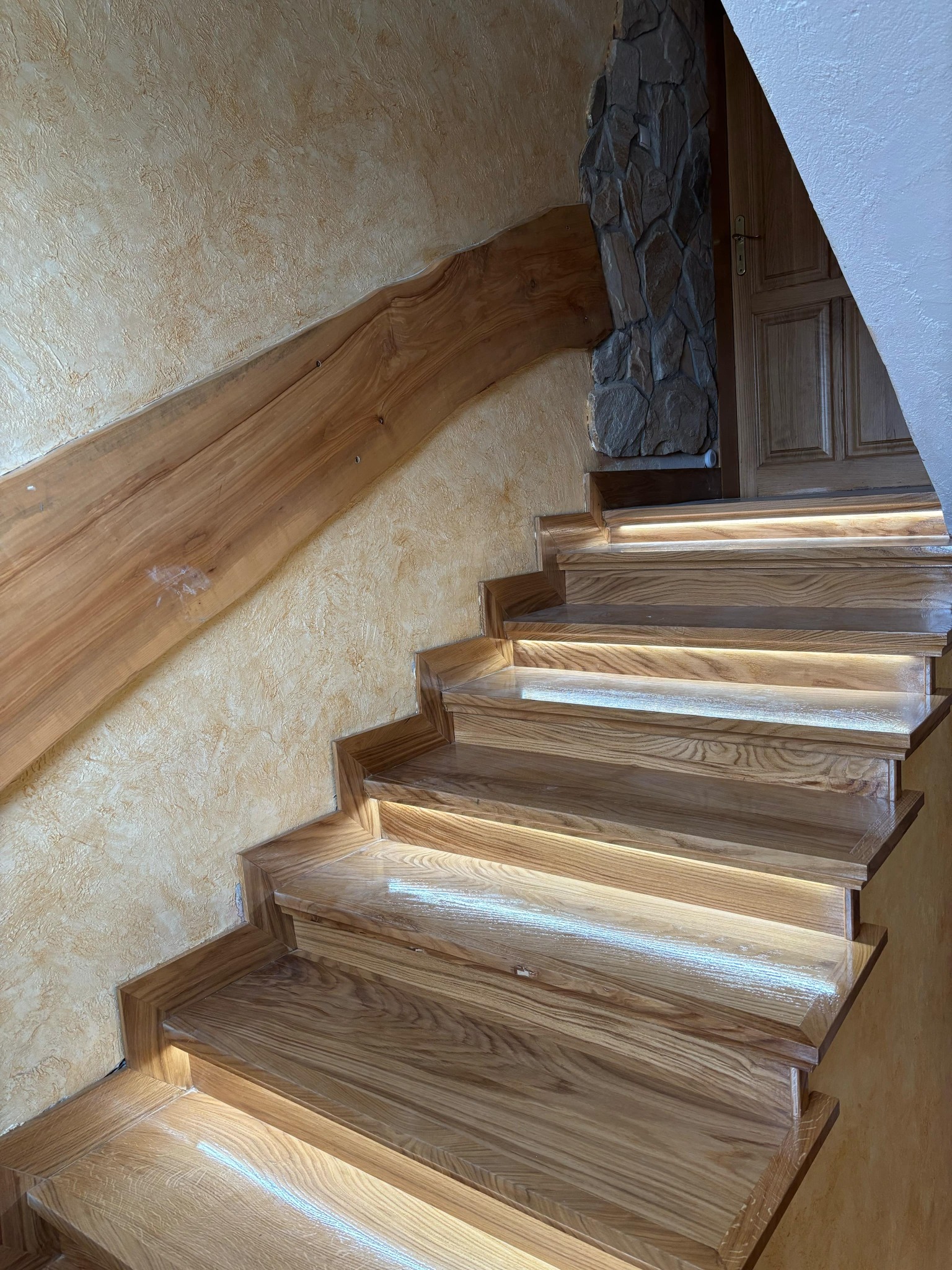 Dębowe schody z podświetleniem LED, drewniana balustrada i ściana o fakturze piaskowca, widok z dołu.