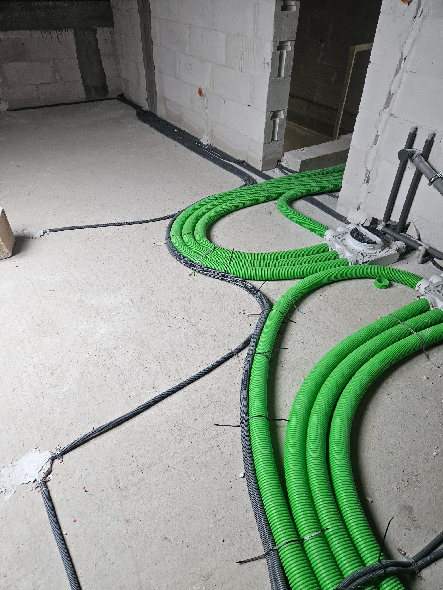 Wnętrze w trakcie instalacji elektrycznej: wiązki zielonych i szarych rur karbowanych prowadzone po surowym betonowym podłożu w pomieszczeniu z białymi ścianami z bloczków.