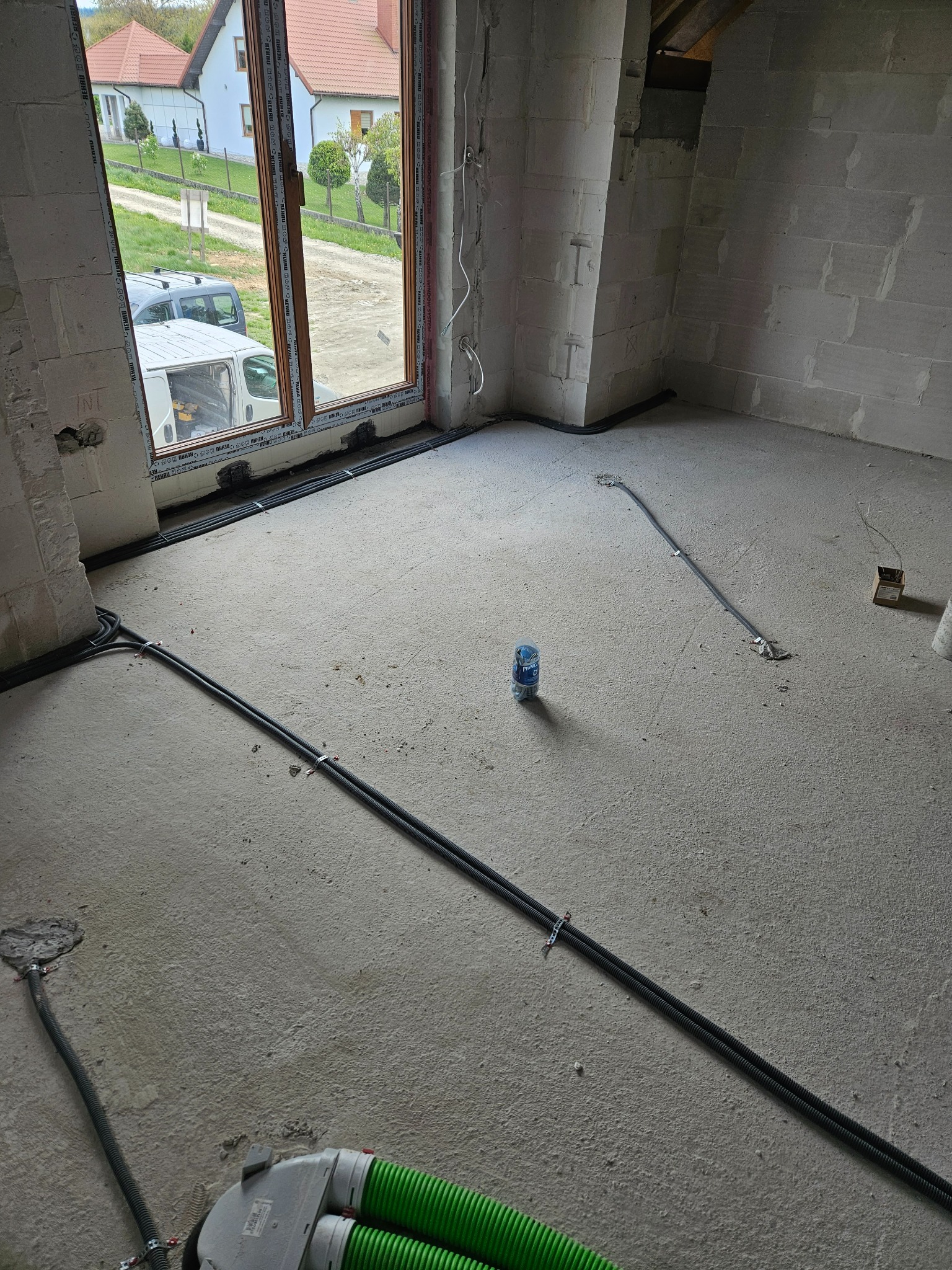 Surowy stan wnętrza w trakcie prac, widoczne kable elektryczne prowadzone po podłodze, przymocowane metalowymi uchwytami, w tle okno z widokiem na domy i samochody, na podłodze butelka wody...