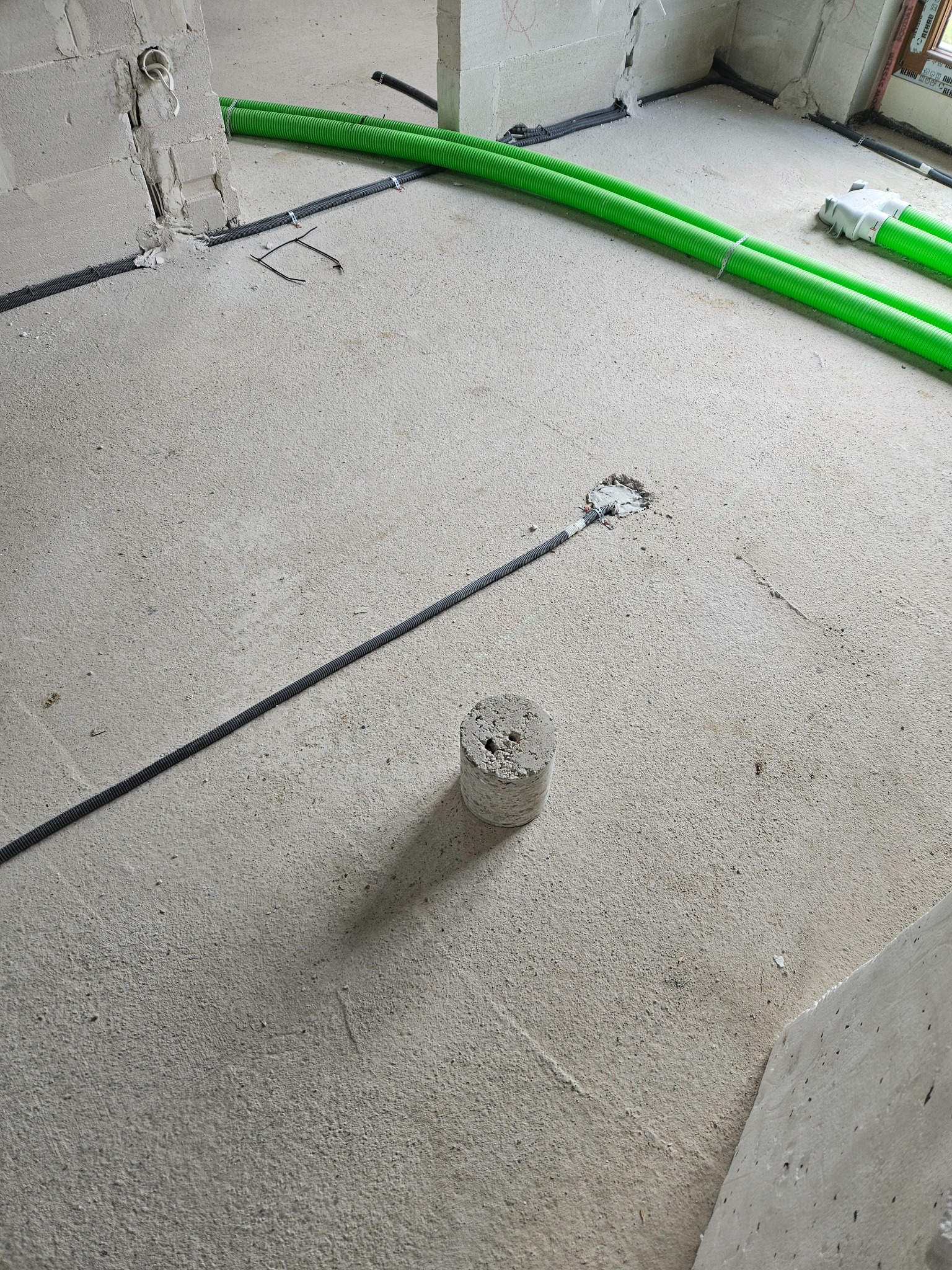 Surowy betonowy podkład z widocznymi zielonymi rurami karbowanymi, czarnymi kablami w peszlu, puszką elektryczną w podłodze i betonowym walcem, etap prac instalacyjnych w nowym budynku.