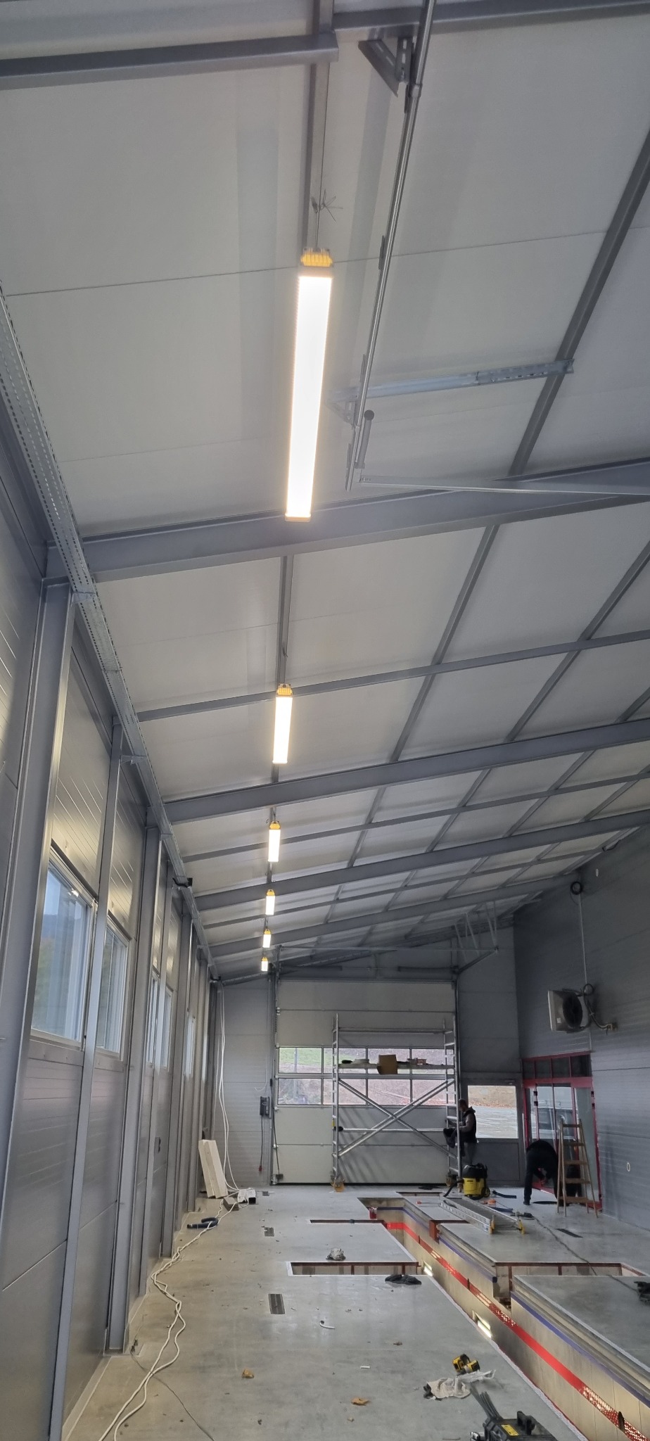 Wnętrze hali warsztatowej z rzędem podłużnych lamp LED zamontowanych pod sufitem, widoczne kanały serwisowe i pracownicy w oddali.
