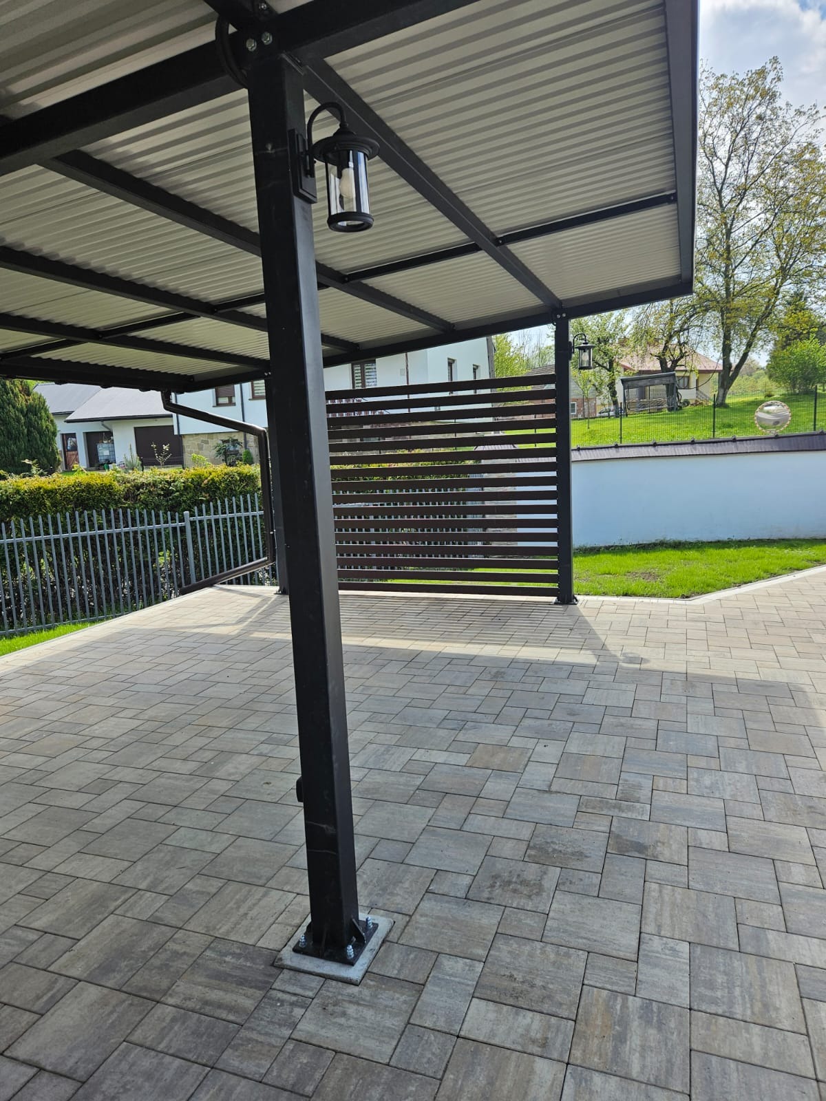 Zadaszenie tarasu z oświetleniem zewnętrznym, widoczne lampy w czarnych obudowach, szara kostka brukowa na podłożu, ogrodzenie oraz zieleń w tle.