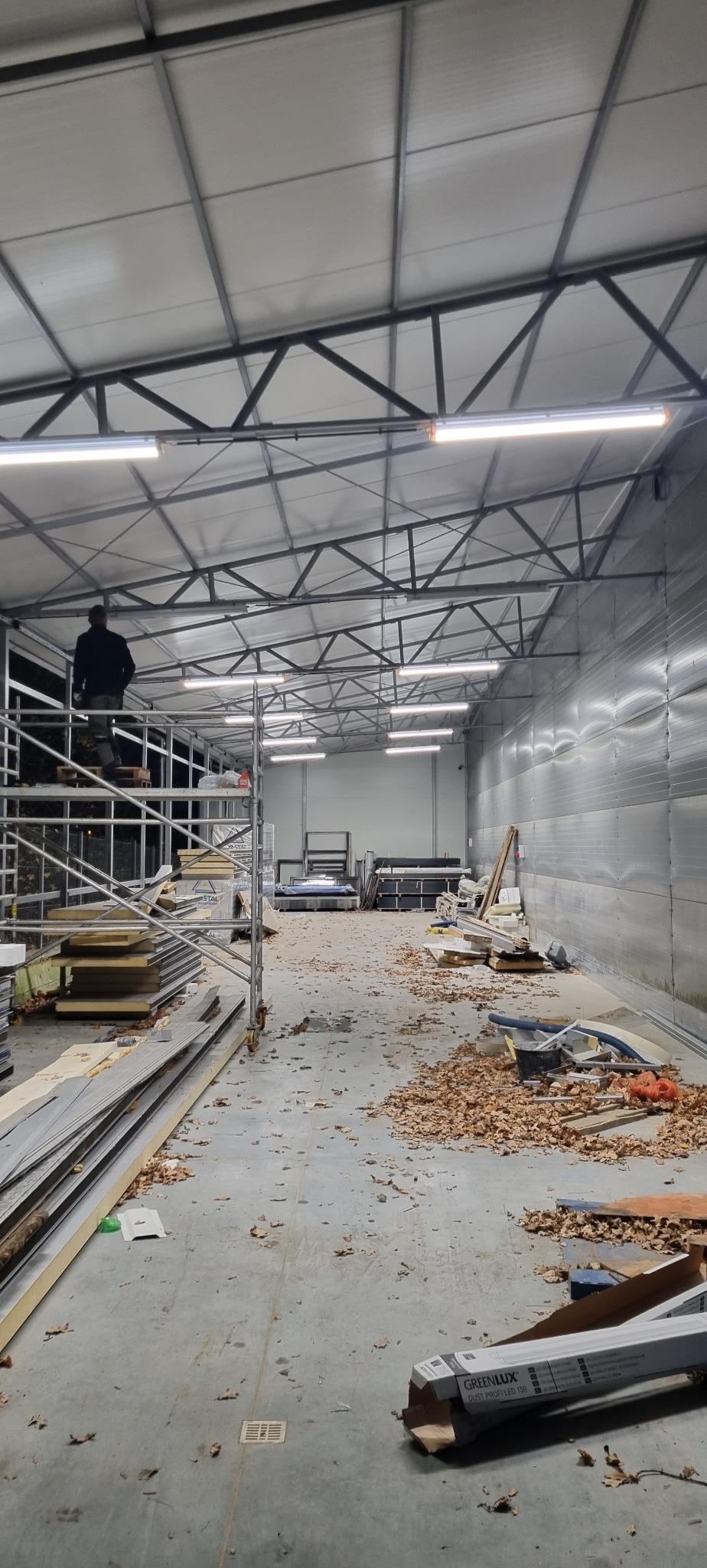 Wnętrze hali w trakcie prac wykończeniowych; widoczne rusztowanie z pracownikiem, zamontowane lampy LED, materiały budowlane i rozsypane liście na podłodze.