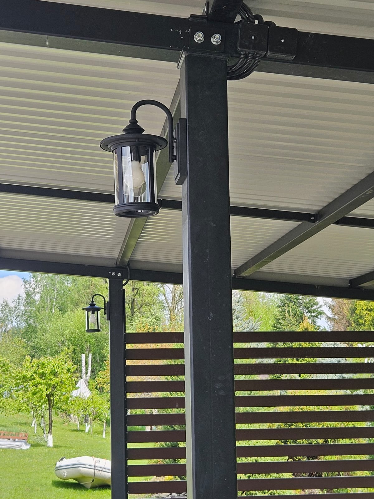 Czarna, zewnętrzna lampa w stylu retro, zamontowana na metalowym słupie pod zadaszeniem, z widocznymi przewodami w peszlu i fragmentem ogrodu w tle.