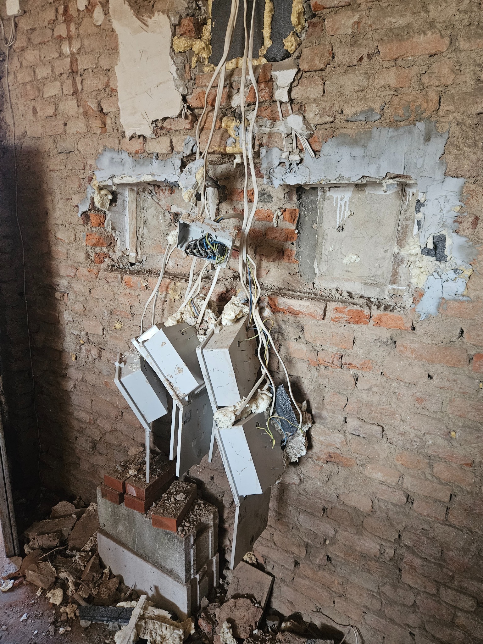 Odsłonięta ceglana ściana w trakcie remontu z wystającymi przewodami elektrycznymi i puszkami podtynkowymi, zabezpieczonymi pianką montażową, gruz na podłodze.
