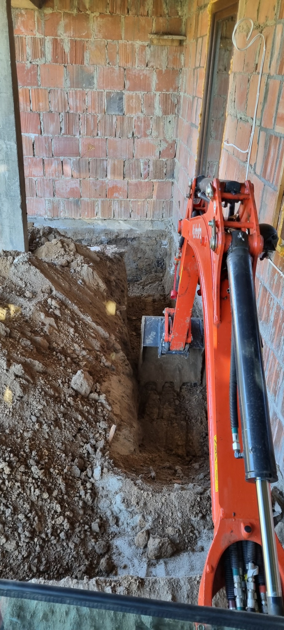 Pomarańczowa mini-koparka Kubota wykonuje wykop w gruncie wewnątrz budynku z surowej cegły, widoczne przewody elektryczne w ścianie.