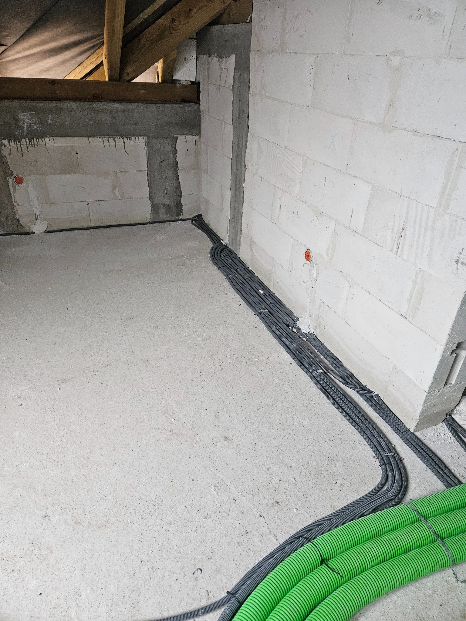 Szare i zielone rury karbowane prowadzone po podłodze w surowym wnętrzu z białych bloczków, widoczne puszki instalacyjne w ścianie i drewniana konstrukcja poddasza.