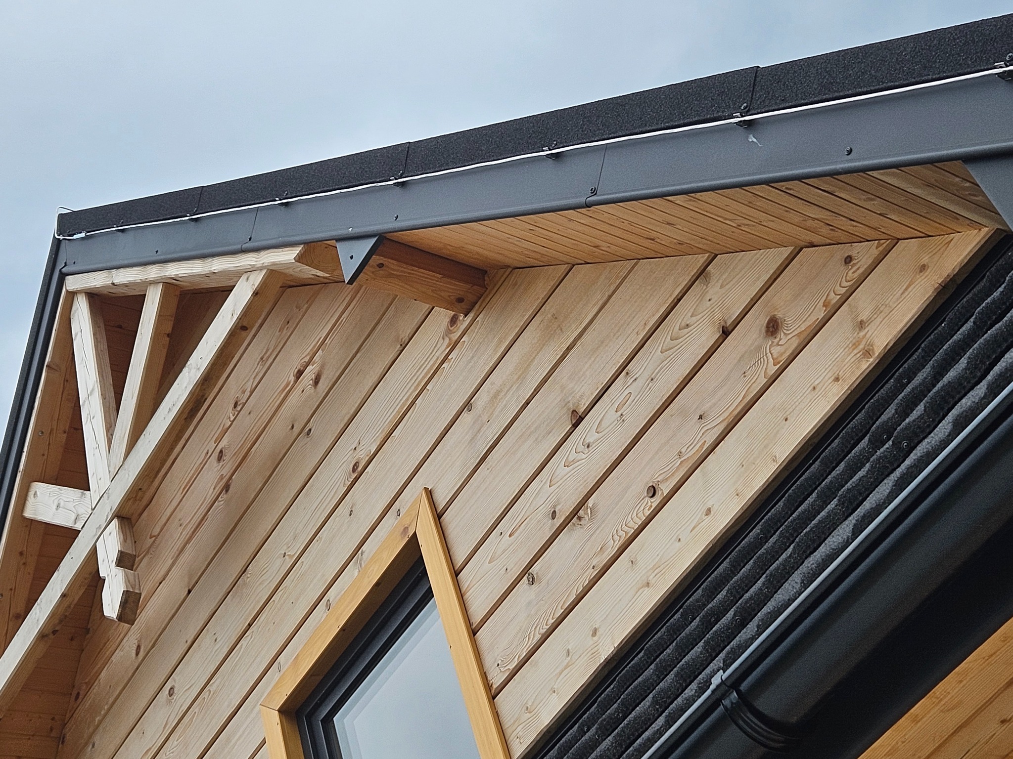 Fragment drewnianej elewacji budynku z widoczną konstrukcją dachu, rynną i oknem o nietypowym kształcie ramy.