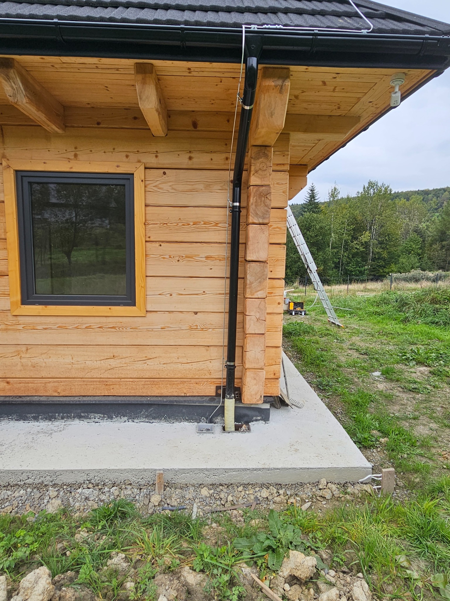 Zewnętrzny widok drewnianego domu z widocznym oknem, rurą spustową i elementami instalacji elektrycznej wychodzącymi z fundamentu, na tle zieleni i drabiny oparta o ścianę.