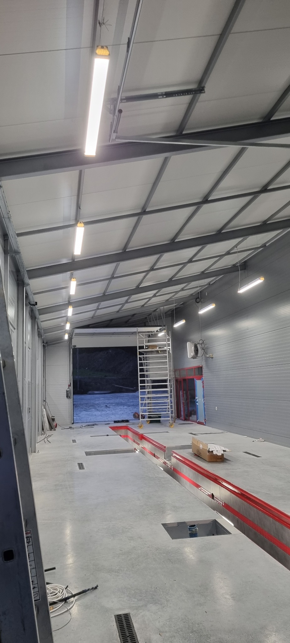 Wnętrze hali warsztatowej z surowym betonem, oświetleniem LED, kanałami naprawczymi z czerwonymi akcentami i rozstawionym rusztowaniem, widoczne okablowanie i narzędzia.
