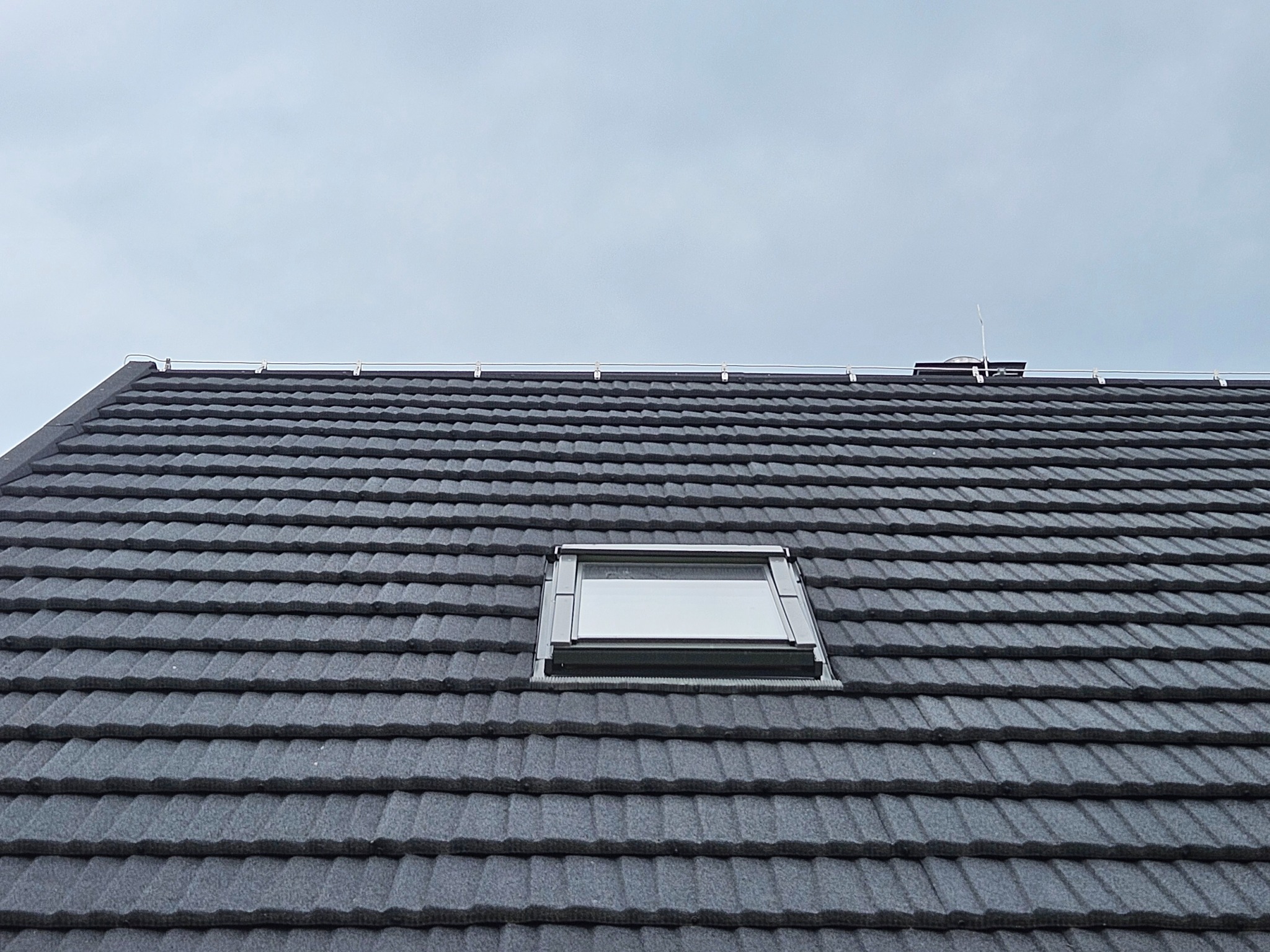 Szary dach z dachówki bitumicznej z zamontowanym oknem dachowym i zabezpieczeniem odgromowym na tle pochmurnego nieba.