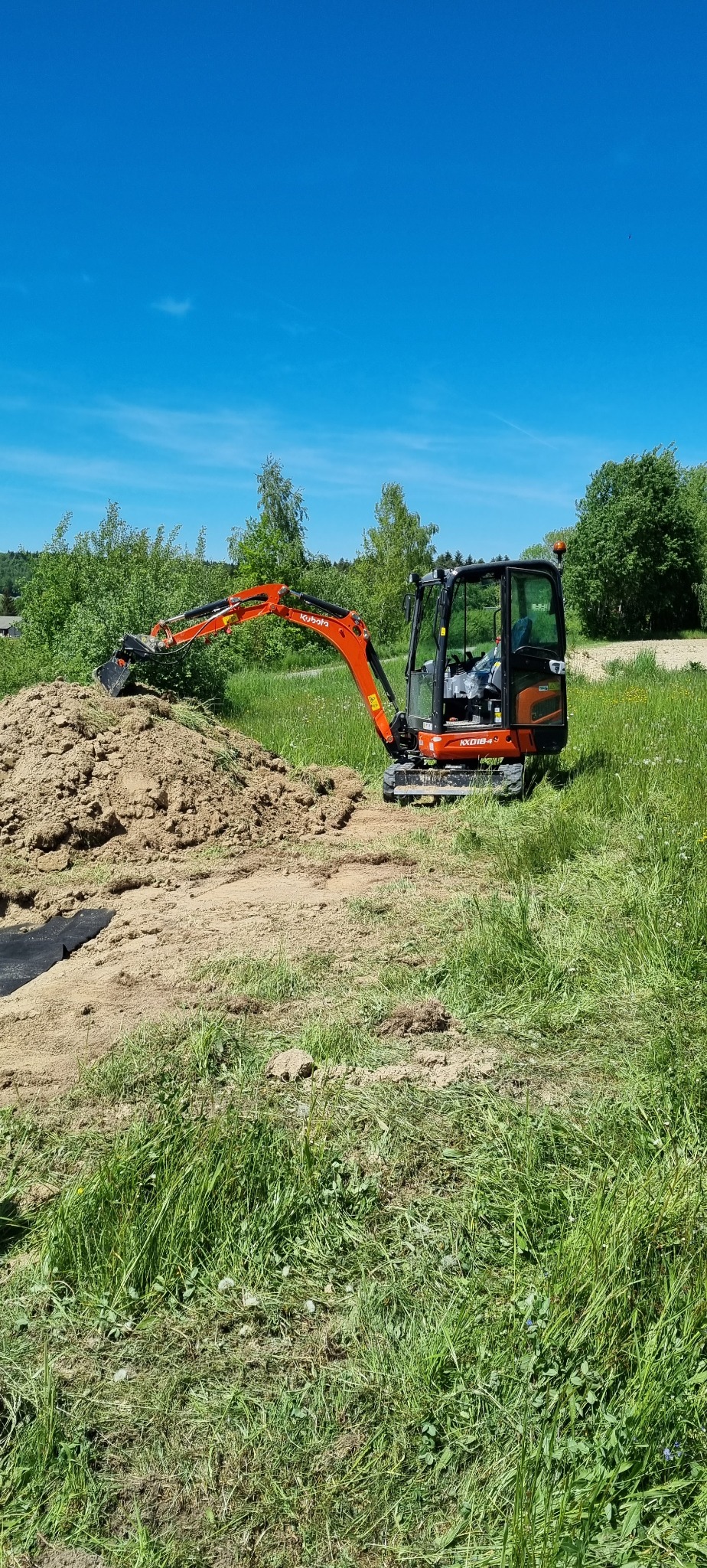 Pomarańczowa mini-koparka Kubota KX018-4 nabiera ziemię z pryzmy na trawiastym terenie w słoneczny dzień.