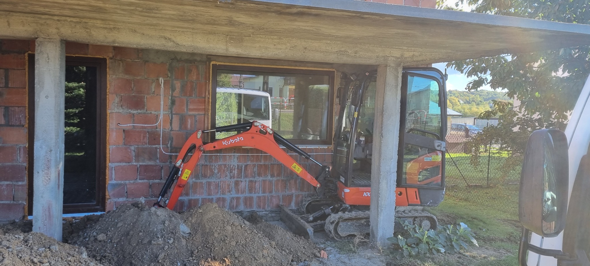 Pomarańczowa mini koparka Kubota KX-41-3V przygotowuje wykop pod fundament budynku z surowej cegły z widocznymi przewodami elektrycznymi.