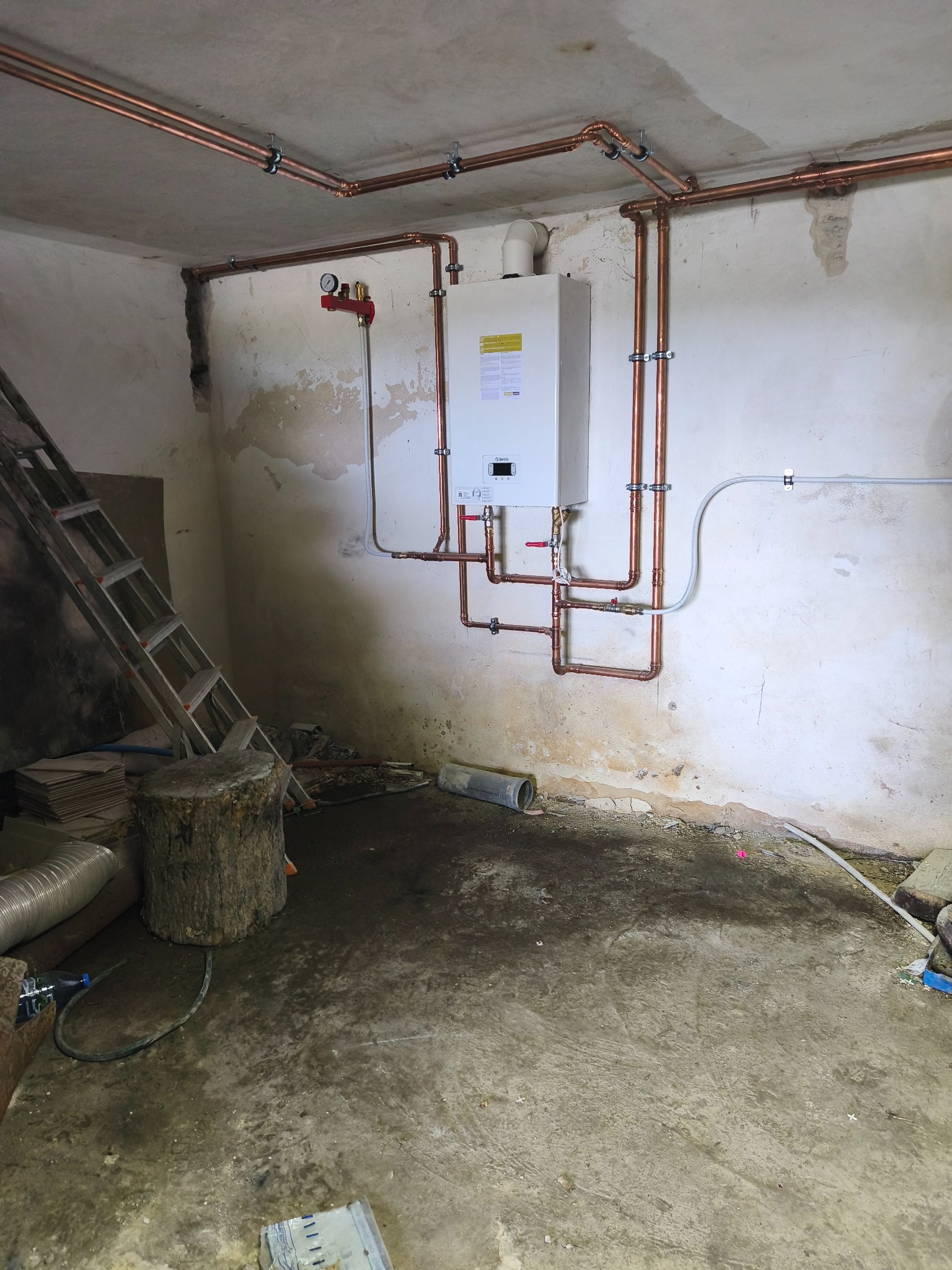 Nowy, biały piec CO zamontowany na ścianie z miedzianymi rurami. Widoczne połączenia i zawory. Surowe wykończenie wnętrza, drabina oparta o ścianę.