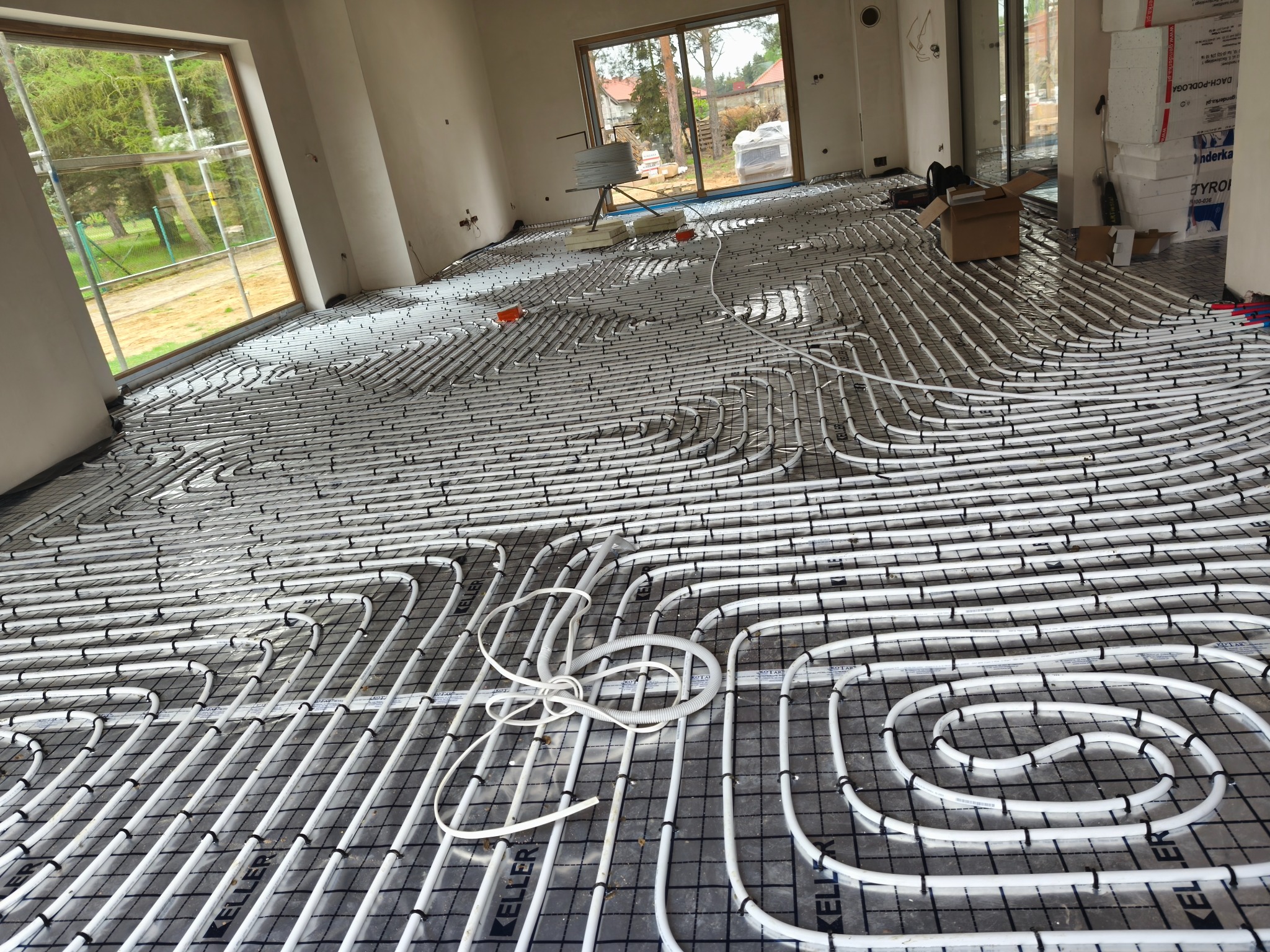 Białe rury ogrzewania podłogowego ułożone na czarnej siatce w jasnym pomieszczeniu z dużymi oknami. Widoczne materiały budowlane i narzędzia.