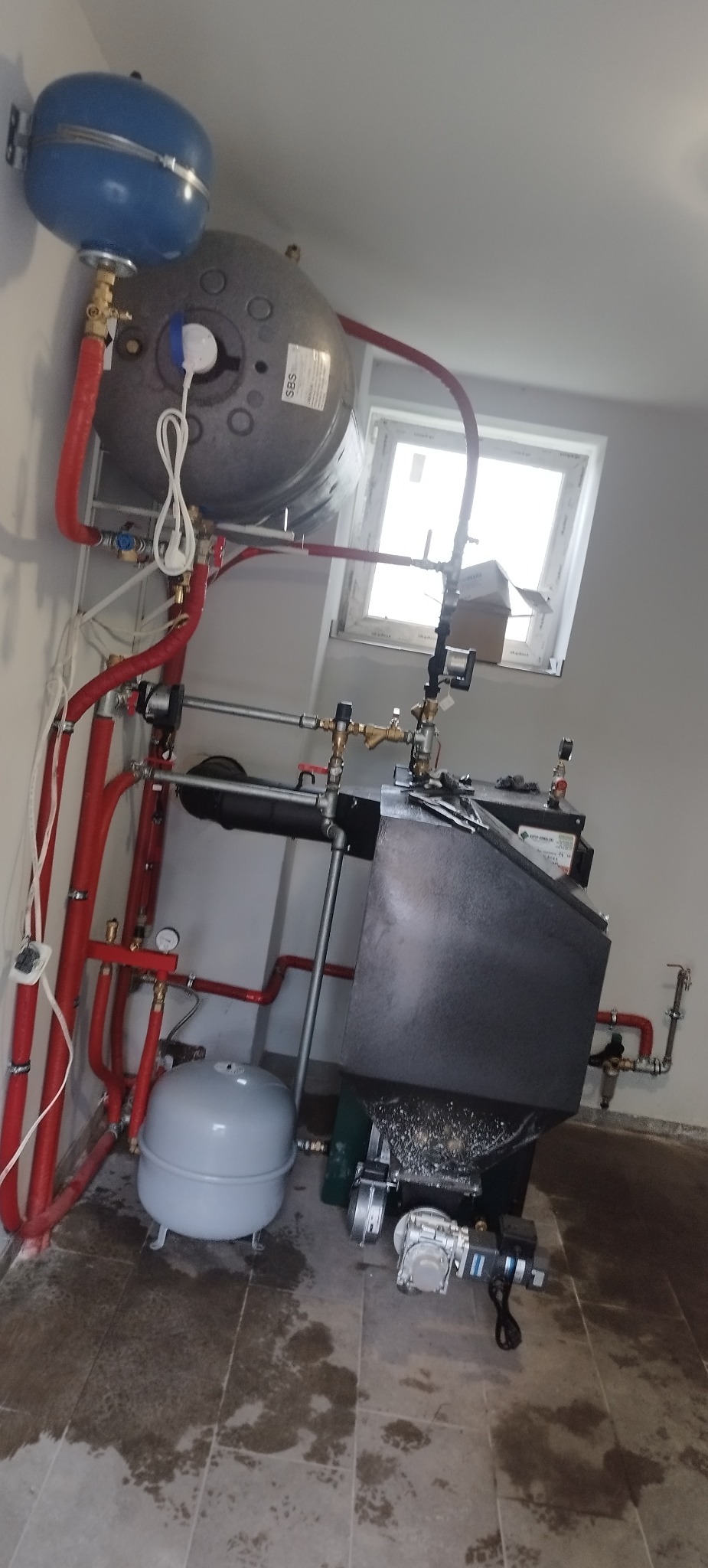 Złożona instalacja grzewcza w kotłowni: piec, zbiorniki wyrównawcze, czerwone rury. Widoczne okno w tle. Elementy połączone, gotowe do użytku.