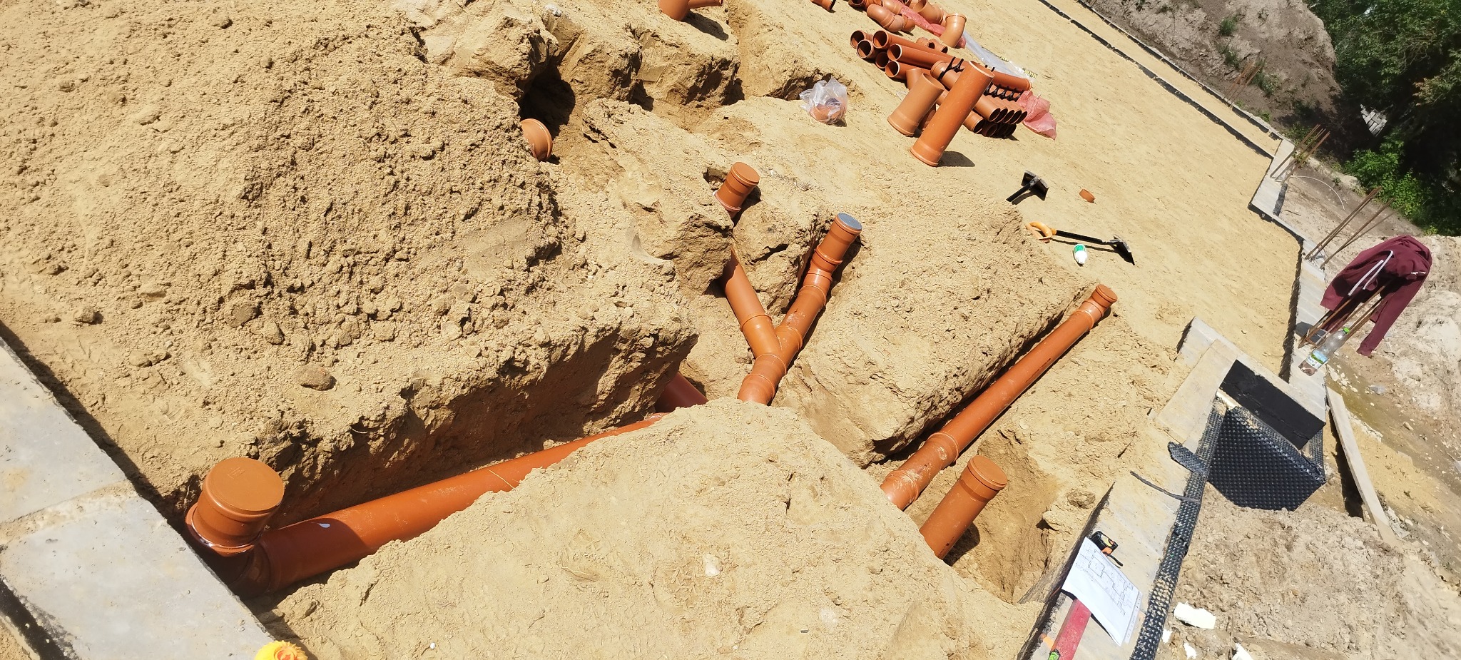 Pomarańczowe rury kanalizacyjne ułożone w wykopie z piasku, widoczne narzędzia i plany. Etap prac przyłącza wod-kan.