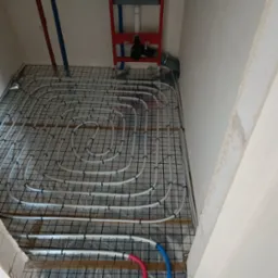 Białe rury ogrzewania podłogowego ułożone na czarnej siatce w surowym wnętrzu łazienki, widoczny stelaż podtynkowy WC.