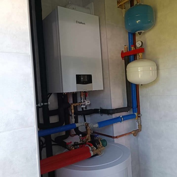 Zamontowany piec gazowy Vaillant z podłączonymi instalacjami, w tym dwoma zbiornikami wyrównawczymi w kolorach niebieskim i białym, oraz licznymi rurami miedzianymi i z tworzywa sztucznego.
