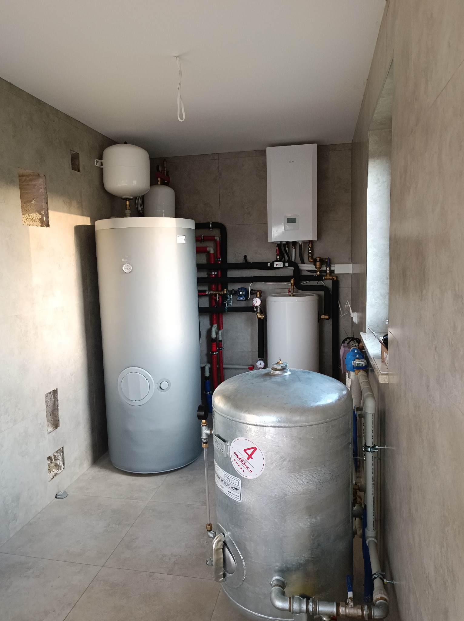 Kotłownia z dwoma zbiornikami akumulacyjnymi, piecem gazowym wiszącym i rozbudowaną instalacją hydrauliczną z rur miedzianych i plastikowych. Na ścianie widoczne niedokończone prace wykończeniowe.