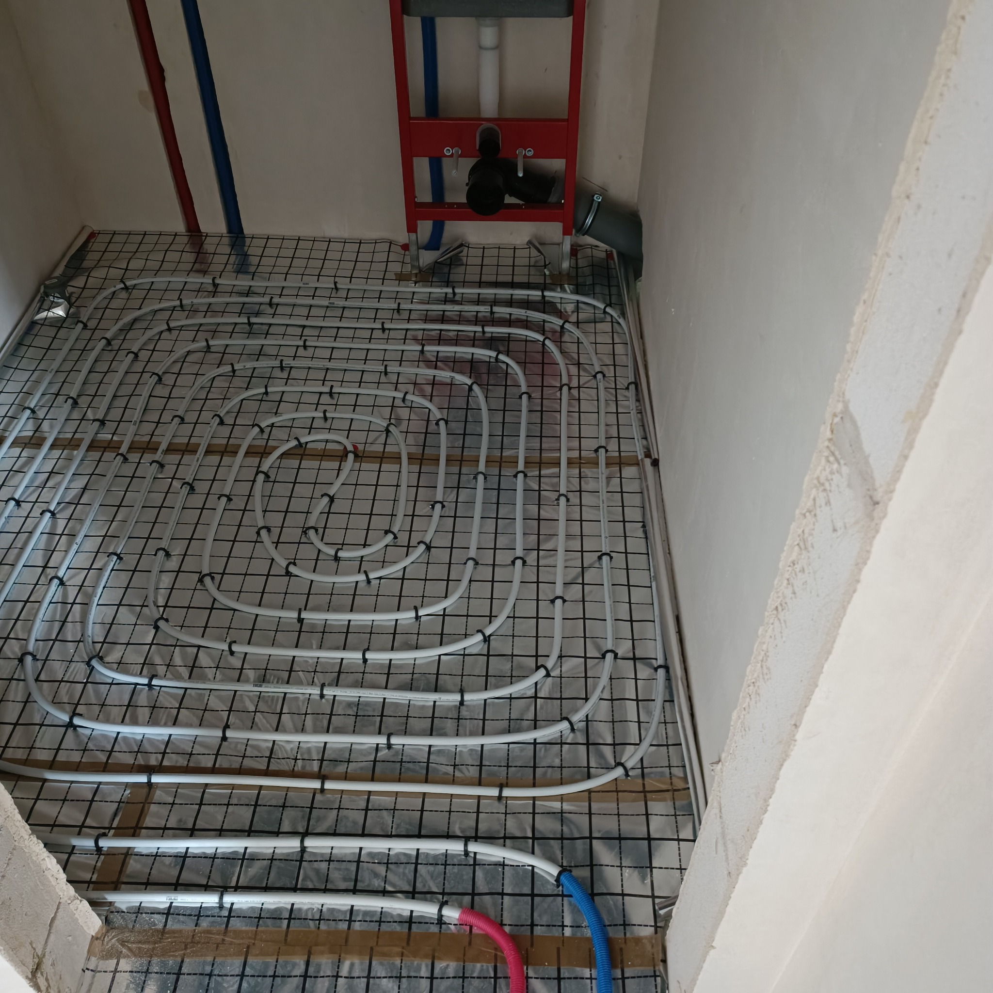 Białe rury ogrzewania podłogowego ułożone na czarnej siatce w surowym wnętrzu łazienki, widoczny stelaż podtynkowy WC.