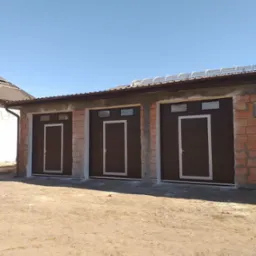 Trzy brązowe segmentowe drzwi garażowe z oknami i jasnymi ramami, zamontowane w surowych murach budynku garażowego w słoneczny dzień.