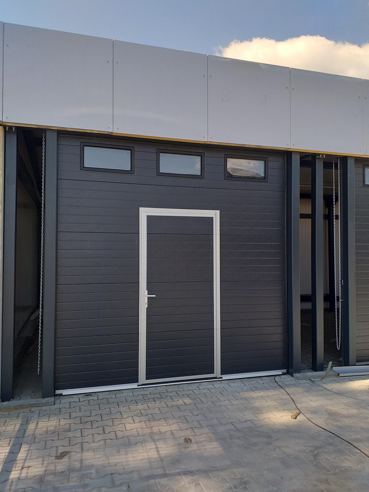 Ciemnoszara brama przemysłowa z trzema małymi oknami u góry oraz drzwiami wejściowymi po prawej stronie, widoczne łańcuchy mechanizmu podnoszenia, na zewnątrz budynku z szarymi panelami.