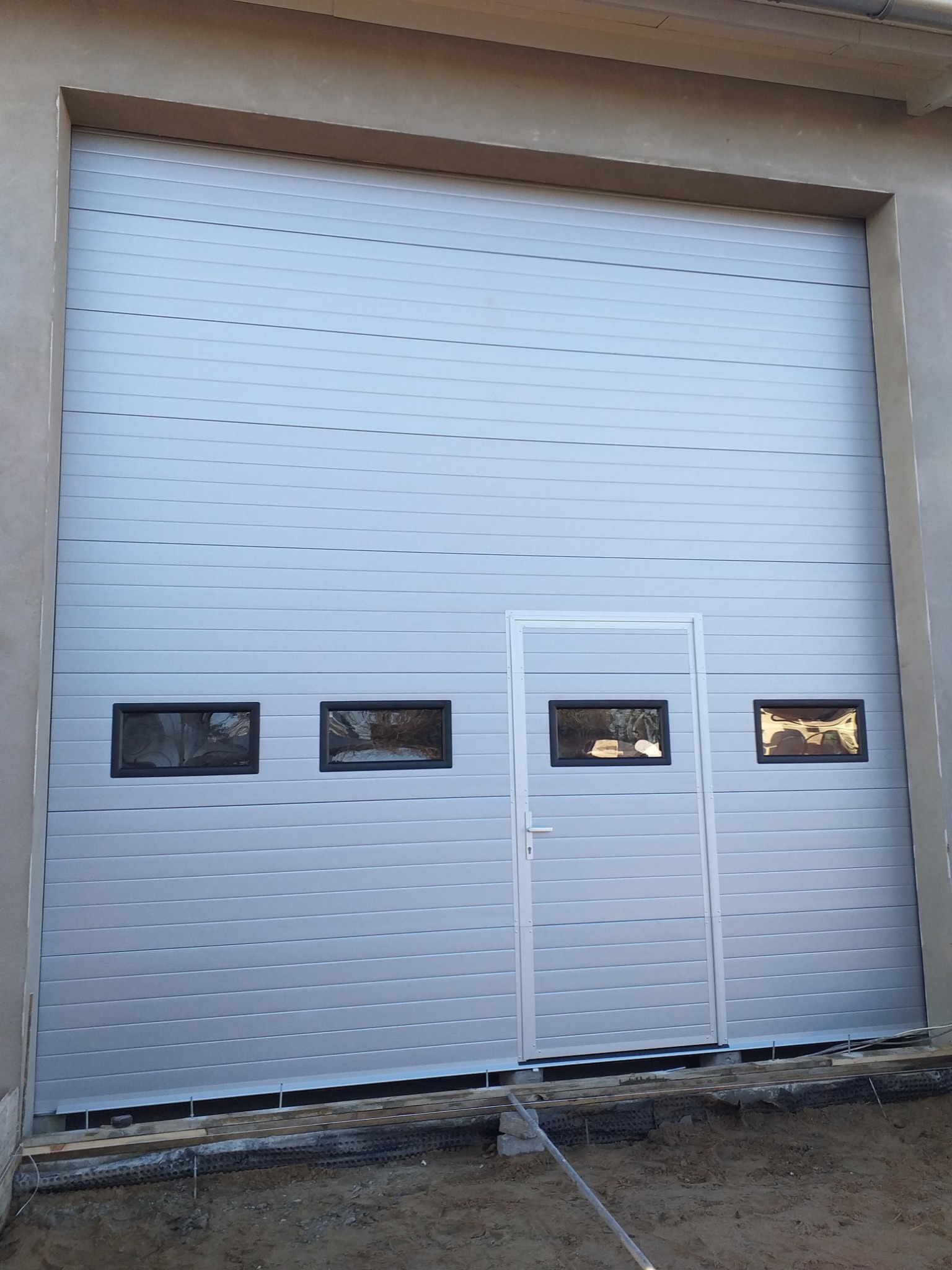 Szara segmentowa brama z wbudowanymi oknami i drzwiami wejściowymi, widoczne przygotowania do montażu, teren wokół w stanie surowym.