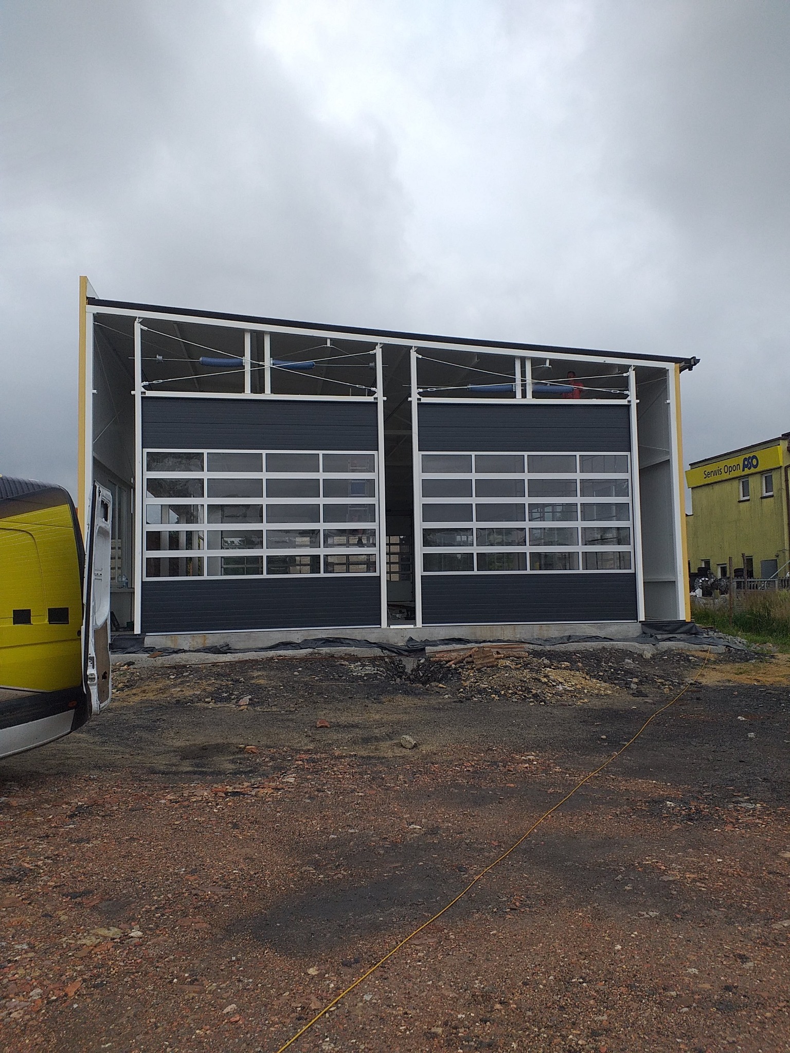 Dwa segmentowe, przeszklone skrzydła bramy przemysłowej w surowym stanie, przed budynkiem z żółtą reklamą serwisu opon.