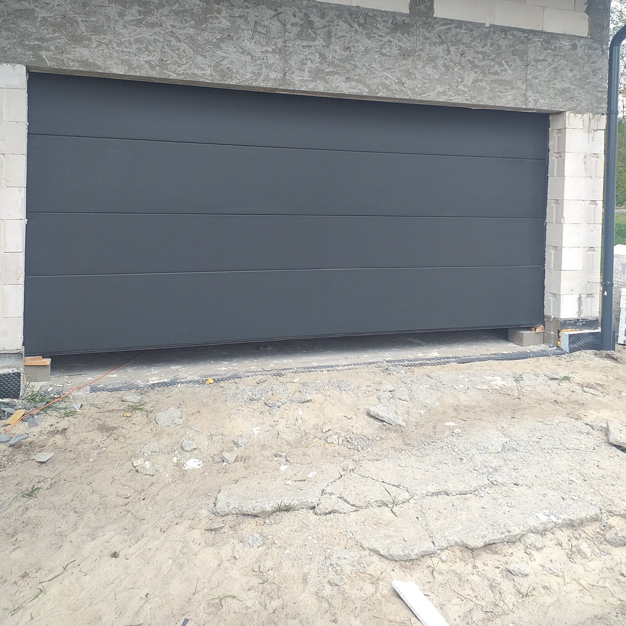 Szara segmentowa brama garażowa w trakcie montażu, widoczne bloczki betonowe i piasek na posadzce.