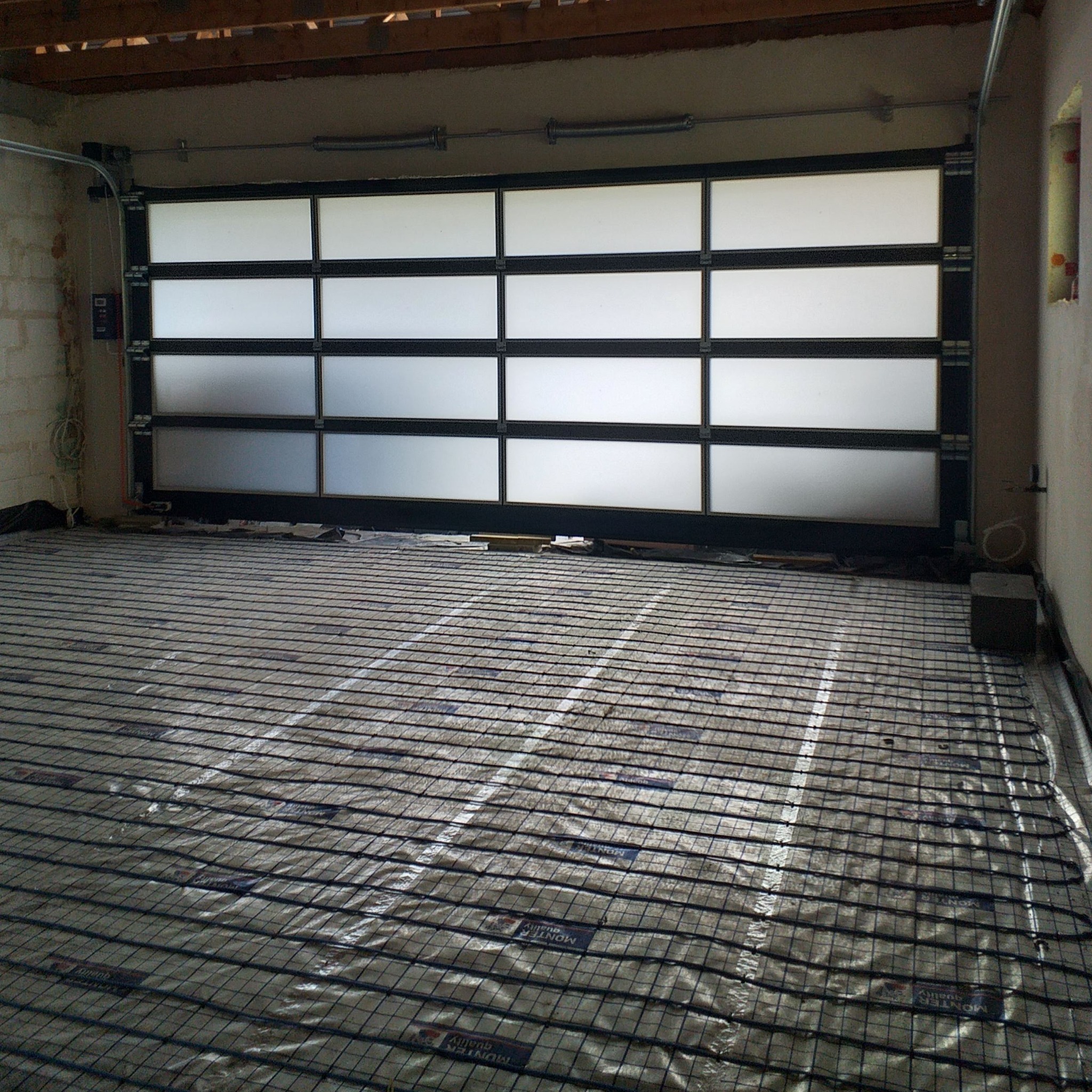 Wnętrze garażu z widoczną sekcyjną bramą z matowego szkła i czarną ramą oraz przygotowanym podłożem pod wylewkę z siatką zbrojeniową i folią.