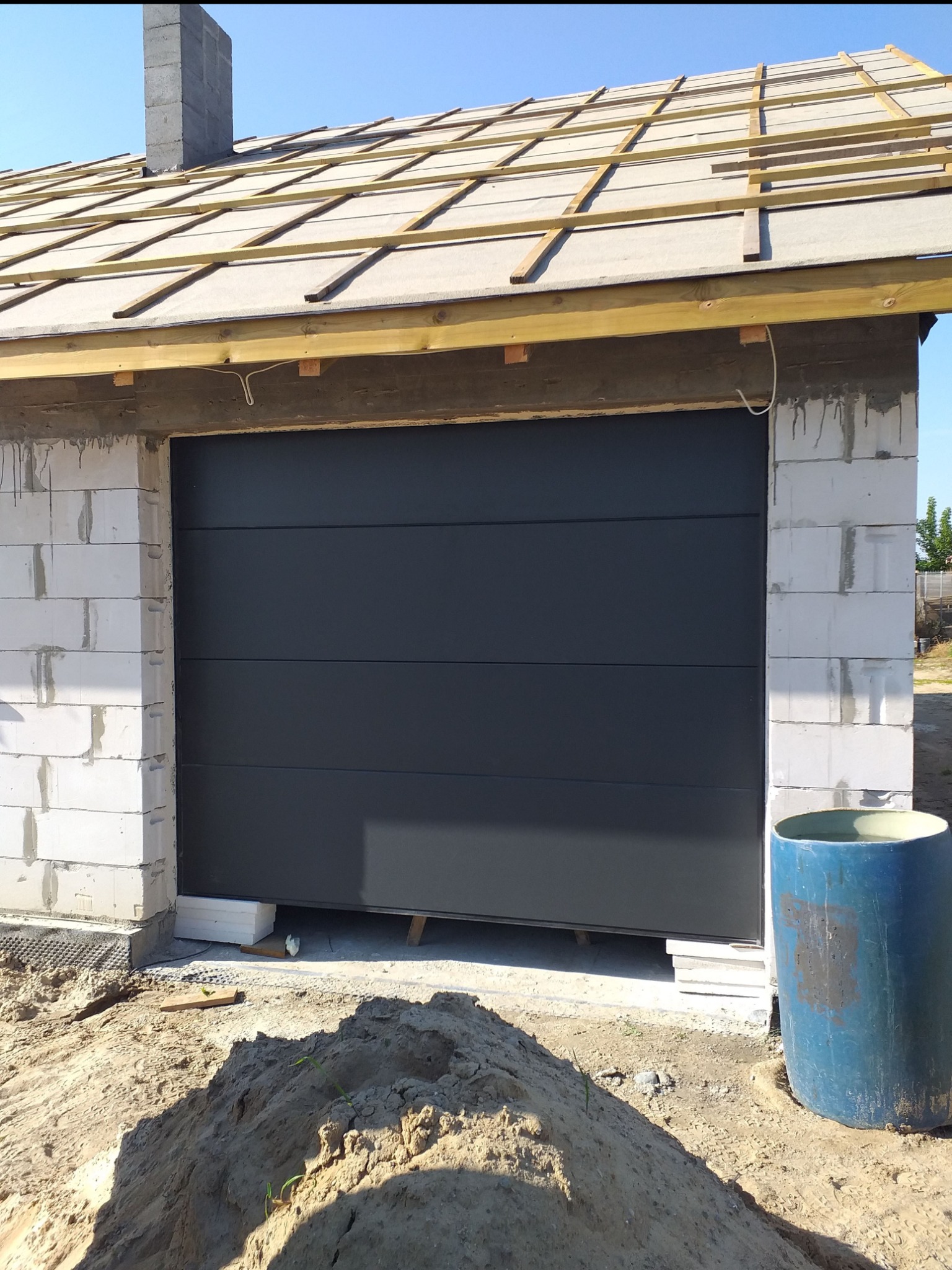 Nowa, antracytowa brama segmentowa w surowym garażu z białego bloczka, widoczne drewniane elementy konstrukcji dachu i sterta piasku przed wjazdem.